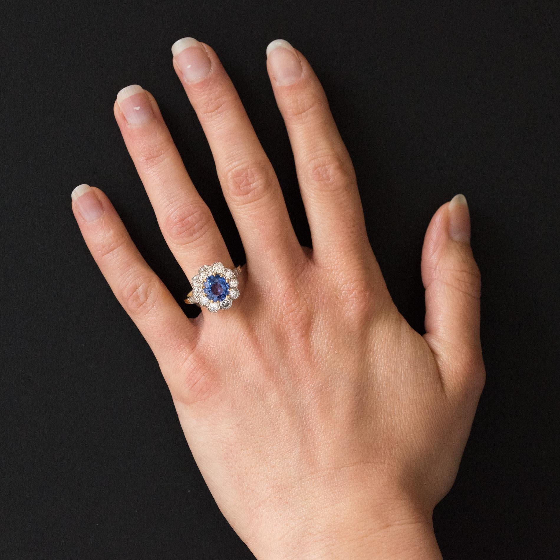 Ring in 18 karat yellow gold and platinum, eagle and dog heads hallmarks.
Modern daisy ring, it is composed of a slightly oval blue sapphire set with claws and surrounded by 10 brilliant-cut diamonds in millegrain setting. 2 x 2 diamonds are set on