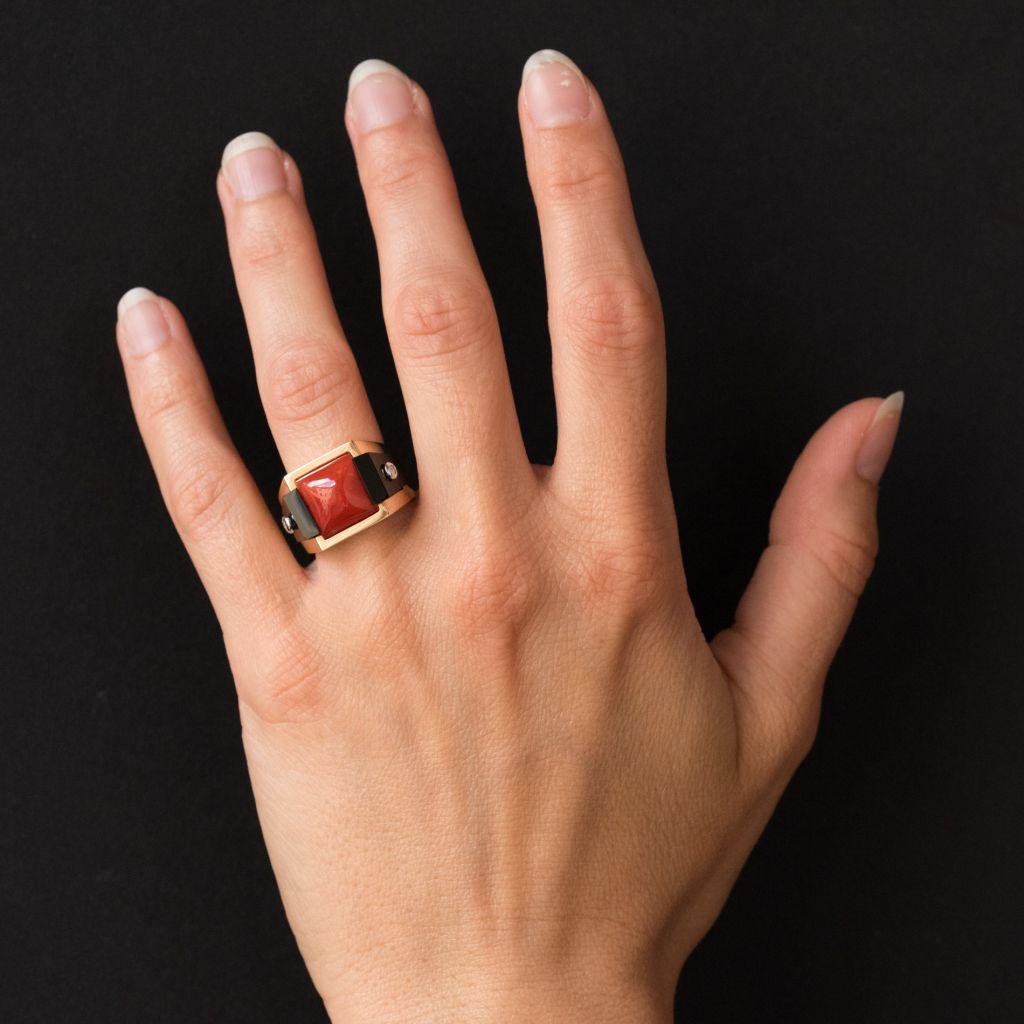 Ring in 18 carats yellow gold.
Yellow gold ring with geometric lines, it is set on its top with a coral carved in sugar loaf supported on either side of an onyx that follows the contours of the ring and set in the center of a brilliant cut