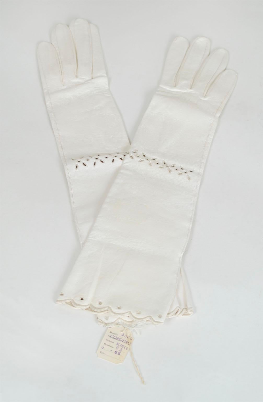 In pristine, brand new condition, these gloves are the pinnacle of femininity thanks to their delicate thread-bound eyelets and scalloped edges. In hard-to-find bright white kidskin leather, their elbow length makes them perfect for black