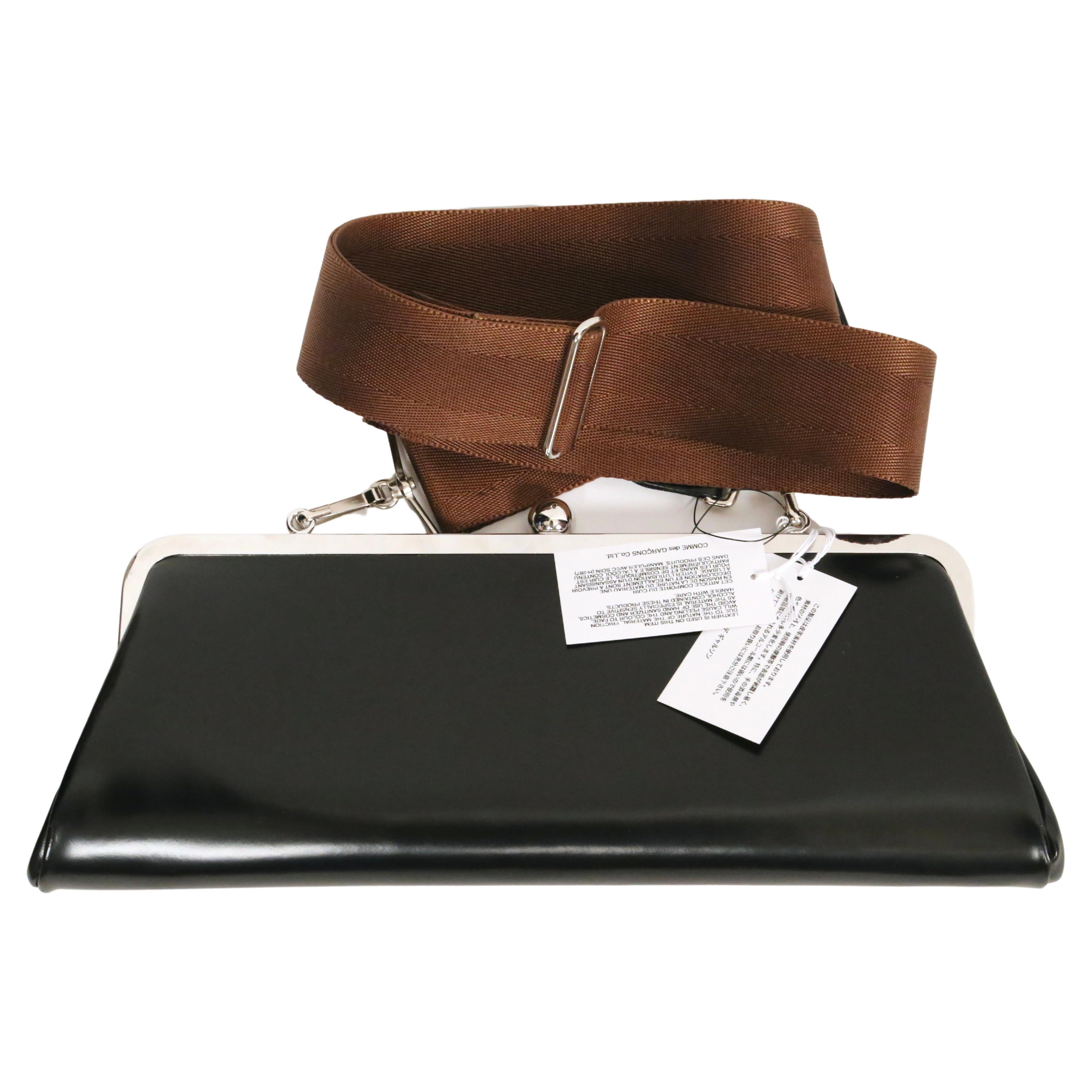 Black, highly polished, leather bag with adjustable brown nylon shoulder strap and silver frame designed by Comme Des Garcons. Approximate measurements: width 14