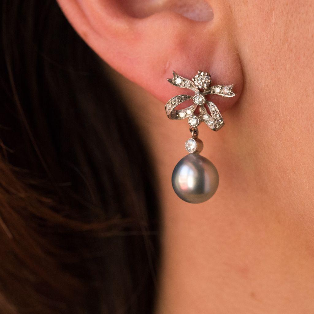 For pierced ears.
Earrings in 18 karat white gold, eagle head hallmark. 
Each grey pearl earring is composed of a bow design claw set with bezel set diamonds with beaded edging. Each holds suspended a bezel set brilliant cut diamond in a beaded