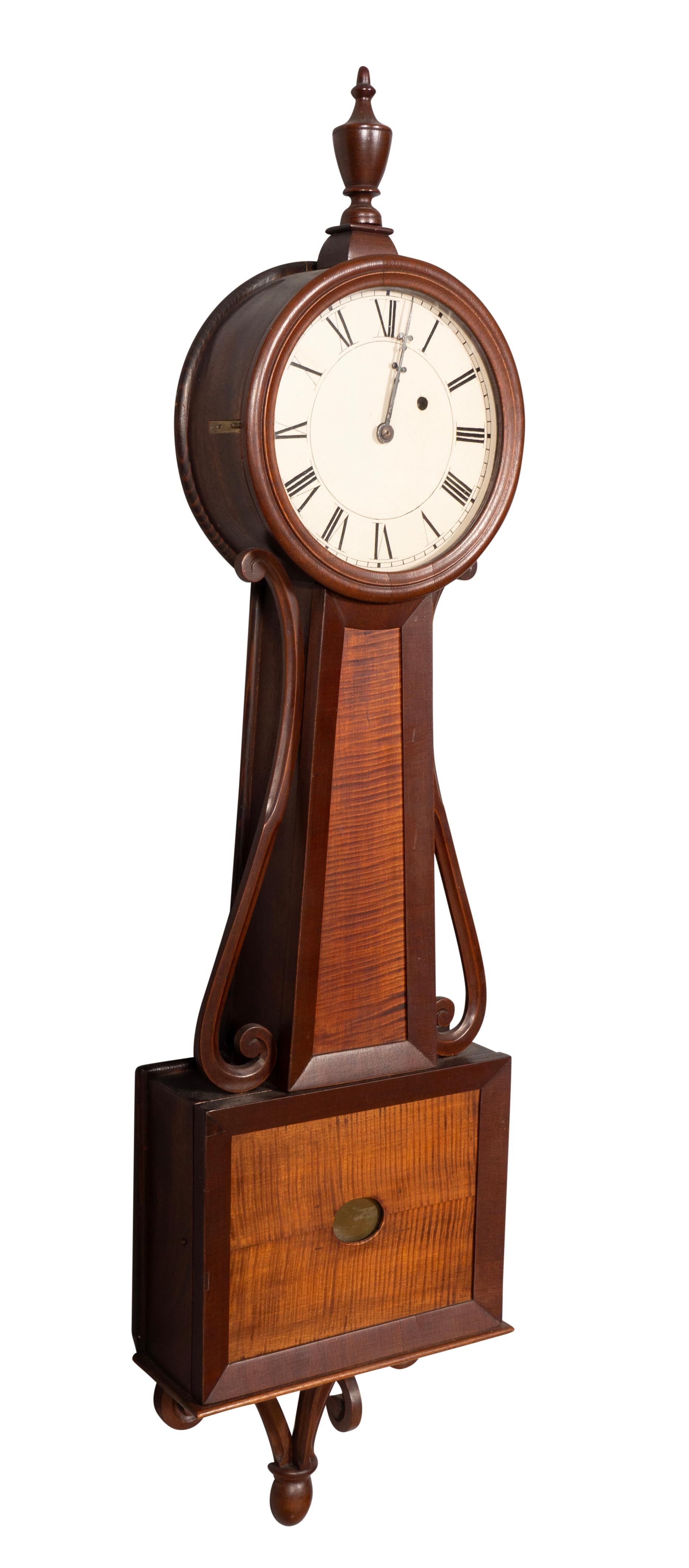 With urn finial over a round clock face with painted iron dial and 
Roman numerals. Hinged glass bezel. Waisted case with finely figured tiger maple flanked by carved mahogany side arms. Conforming door below enclosing the pendulum base visible