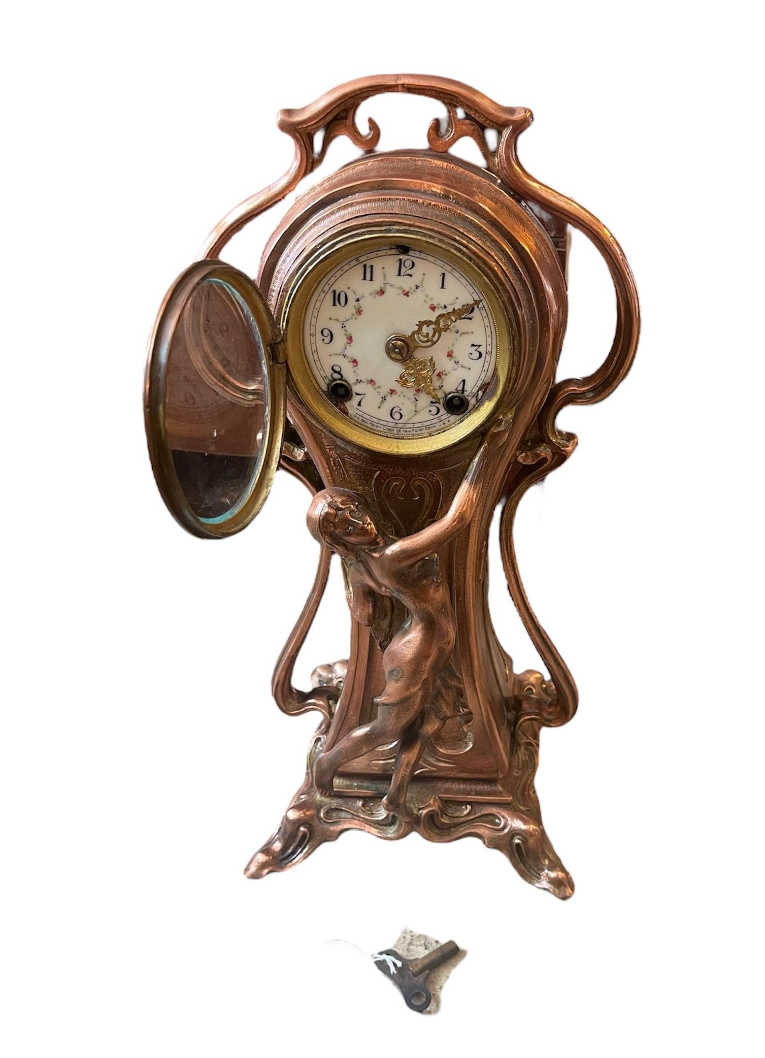 This is an Art Nouveau Style Table Clock. It depicts a small balloon shape clock mounted in copper color metal. A standing young nymph is adorning the front of the case while holding its face. The white enamel round dial of the clock has Arabic