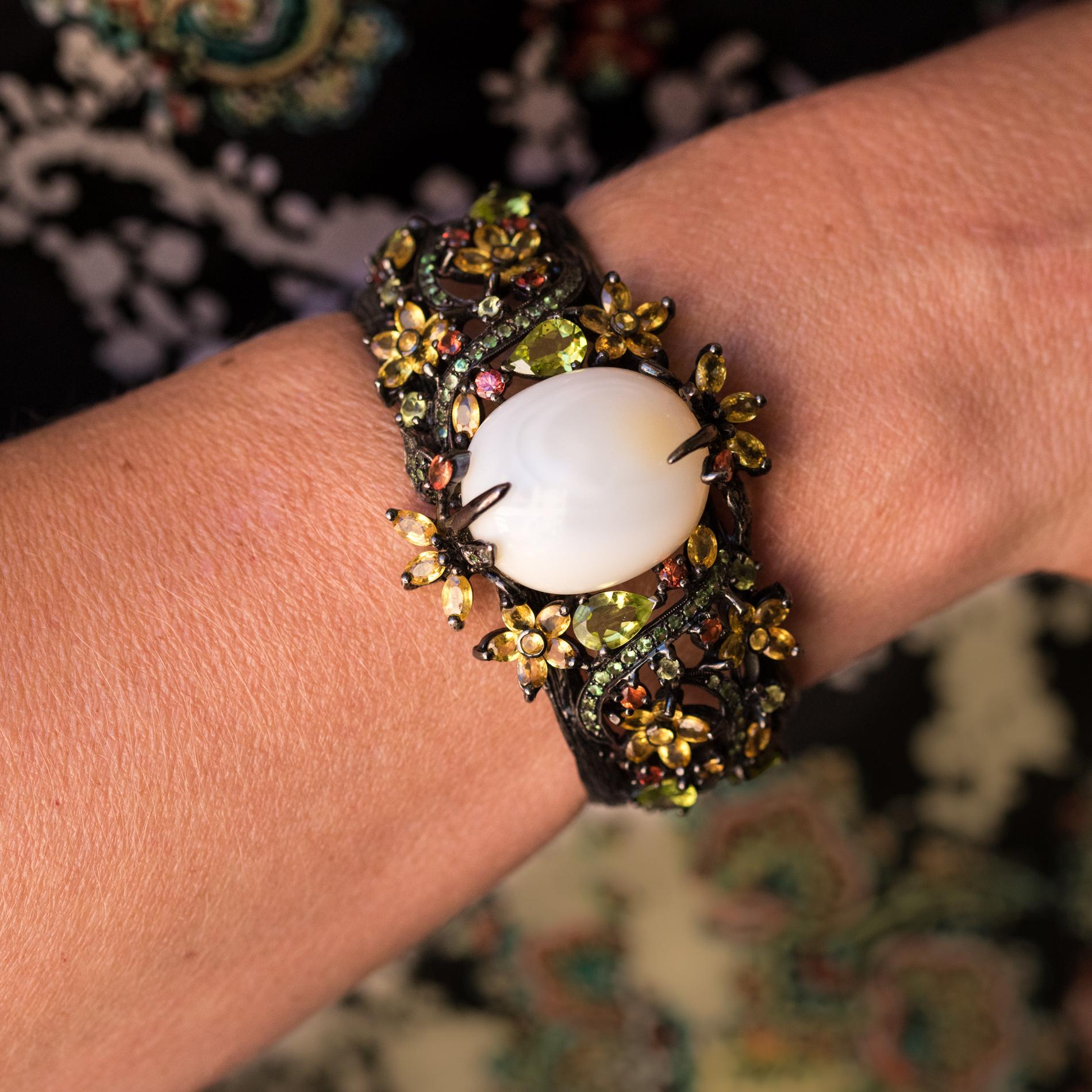 Bracelet in silver, black rhodium.
Impressive cuff, this bracelet is set on its top with a common white cabochon opal. The whole bracelet is openwork and represents a decor of branches, leaves and flowers set with peridots, green tsavorite garnets,