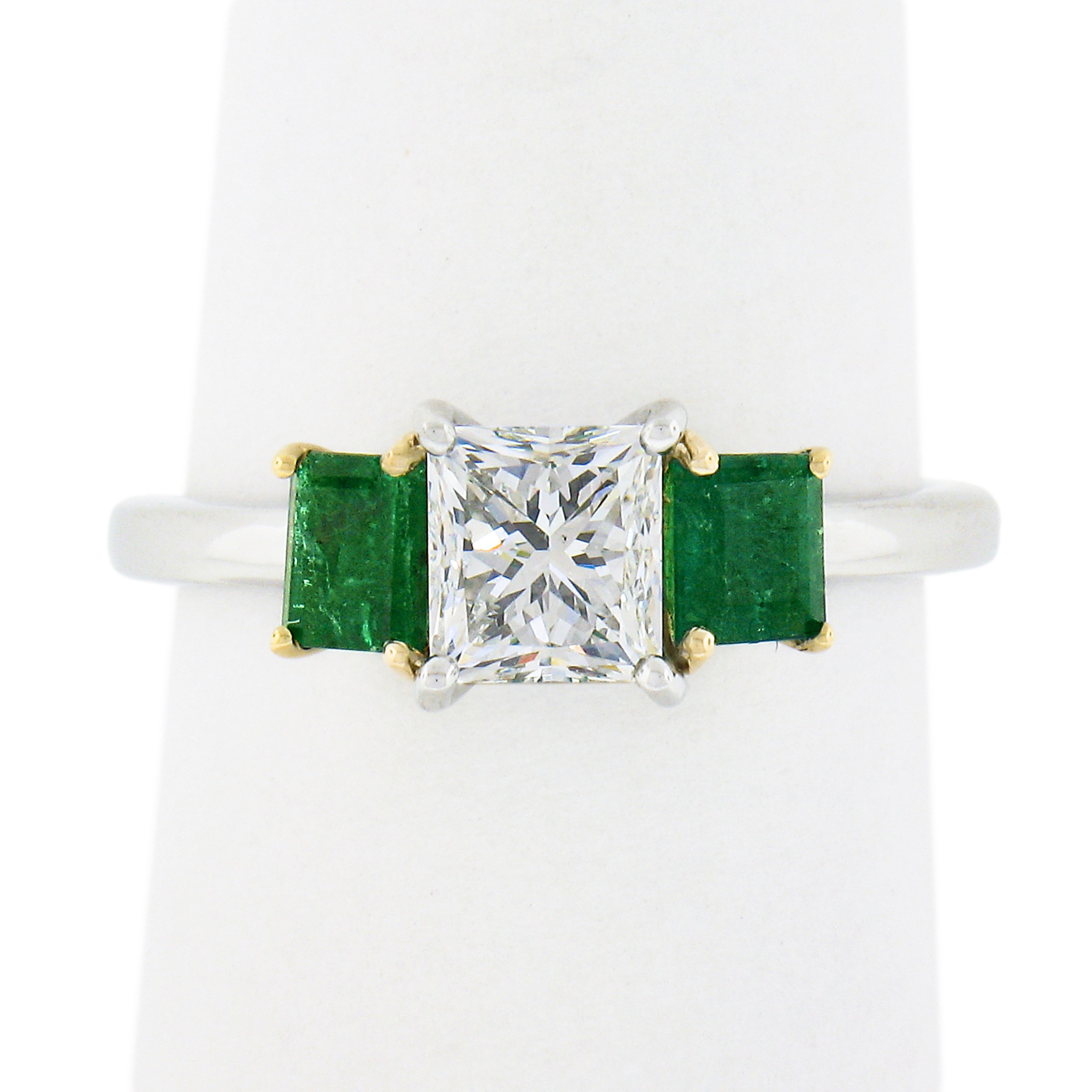 This elegant, brand new and custom designed diamond and emerald three stone engagement ring is crafted in solid platinum and 18k yellow gold. It features a truly gorgeous, GIA certified, princess cut diamond solitaire prong set at the center and is