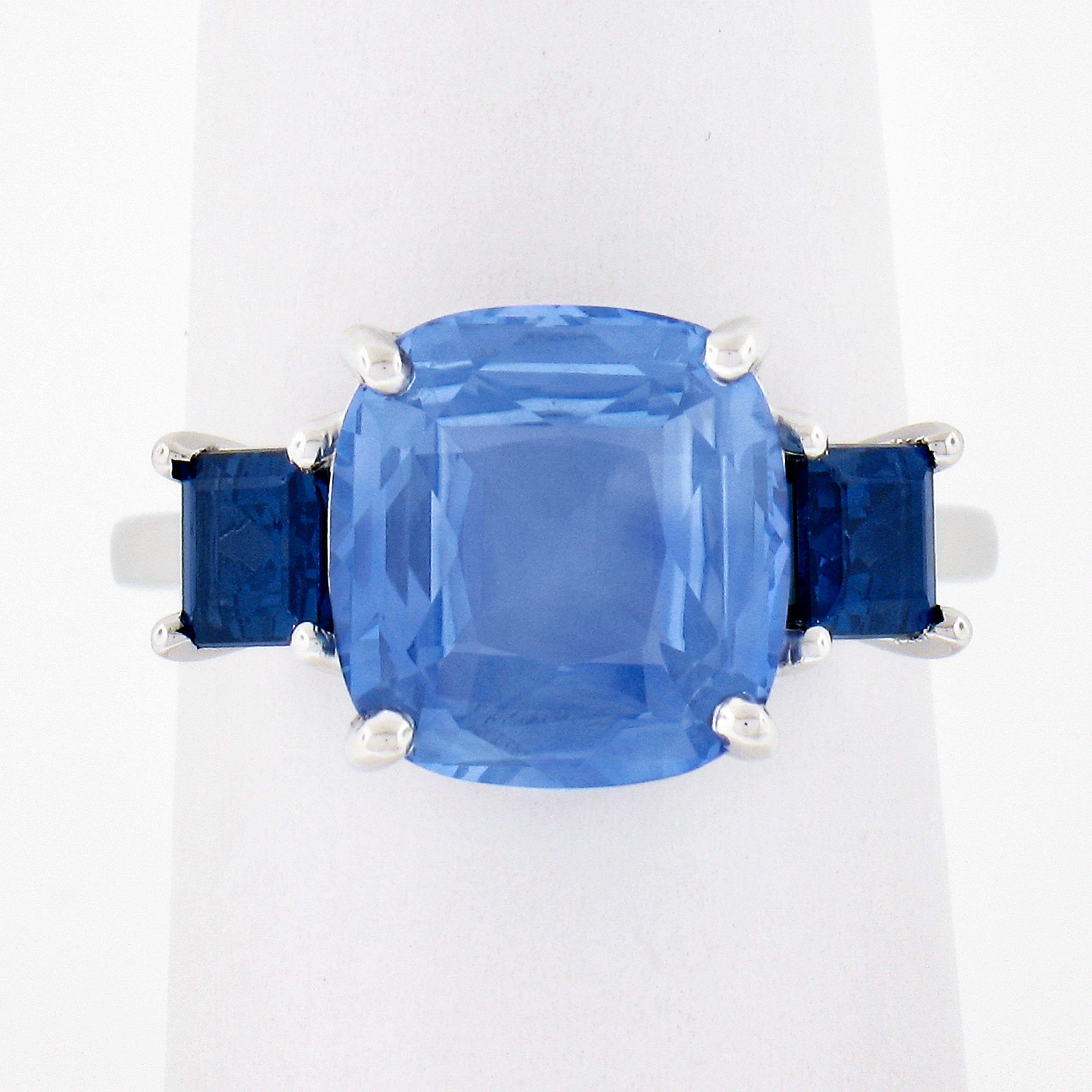 This very elegant sapphire three stone engagement style ring is newly crafted in solid platinum. It features a gorgeous GIA certified, 5.01 carat, cushion cut sapphire neatly prong set at the center showing a beautiful cornflower blue color with