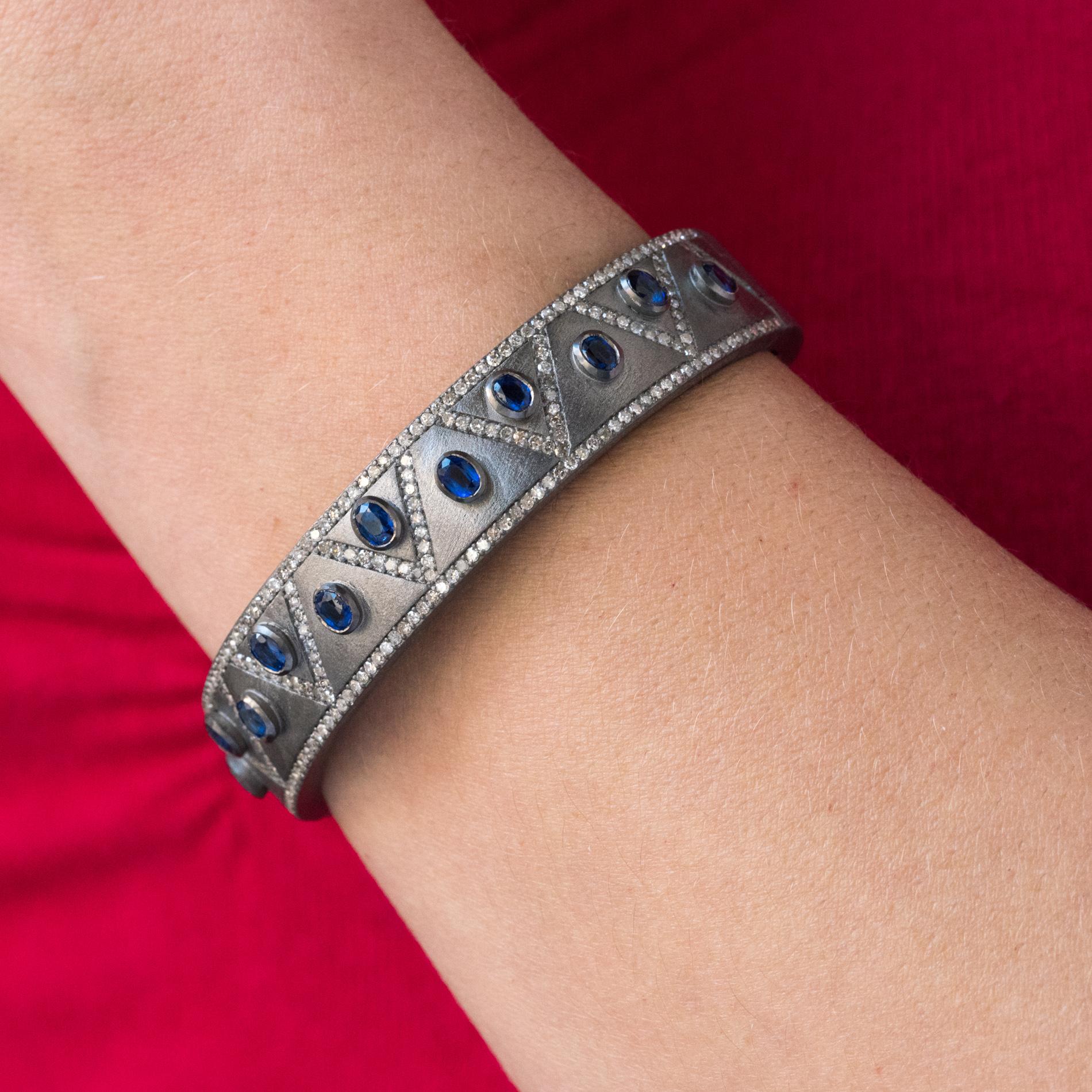 Bracelet en argent, grisé et matifié.
Rigide, ce bracelet en argent est orné sur son dessus de motifs à chevrons sertis de diamants y compris sur les bords. 11 cyanites ovales bleues en serti clos séparent ces motifs. L'intérieur est perforé de