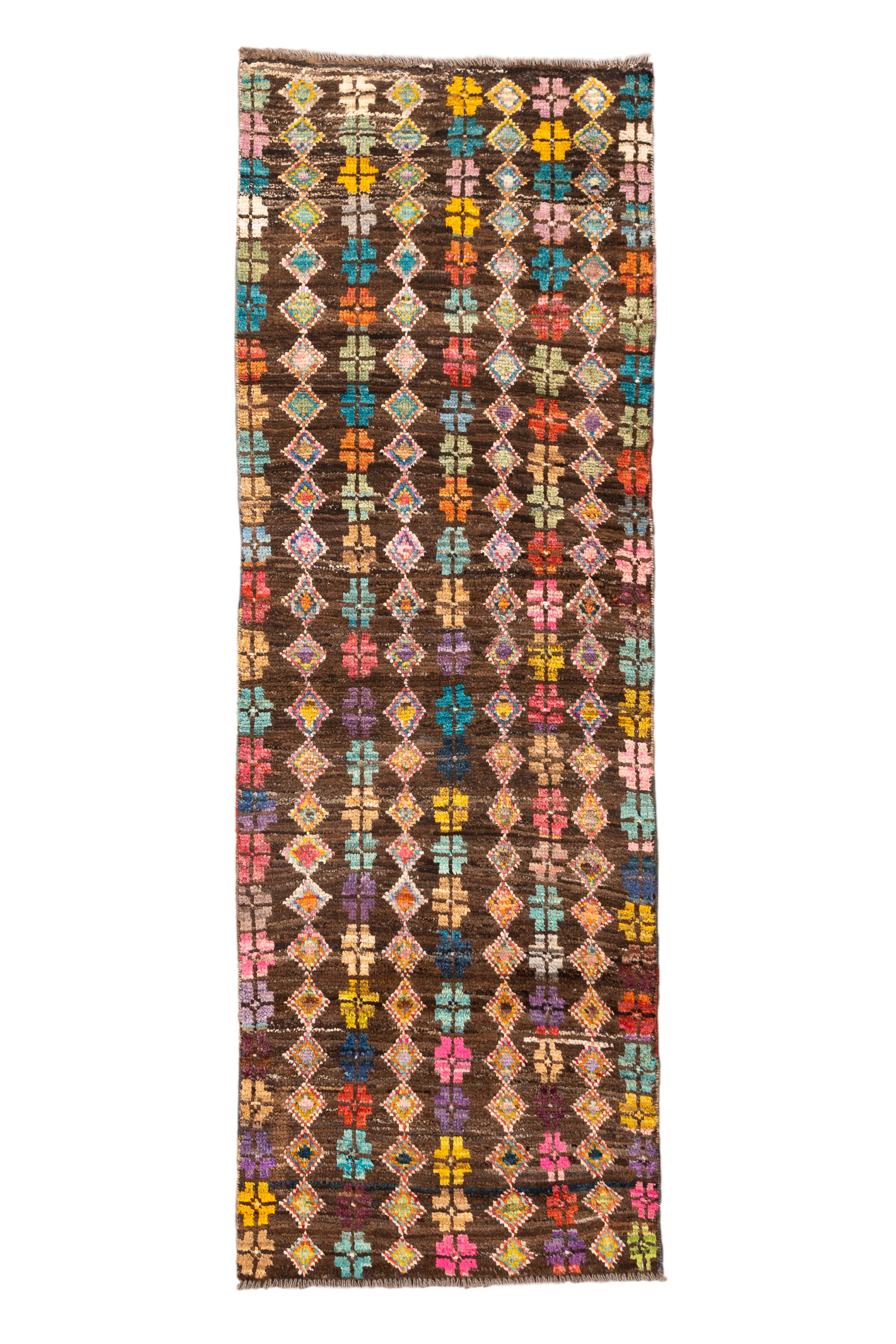  Esta llamativa alfombra Tulu vintage, tejida a mano en Turquía, presenta una disposición rítmica de motivos cruzados en tonos vivos -turquesa, azafrán, lila, rosa, coral y verde azulado- sobre un rico fondo marrón espresso. El estampado pulsa con