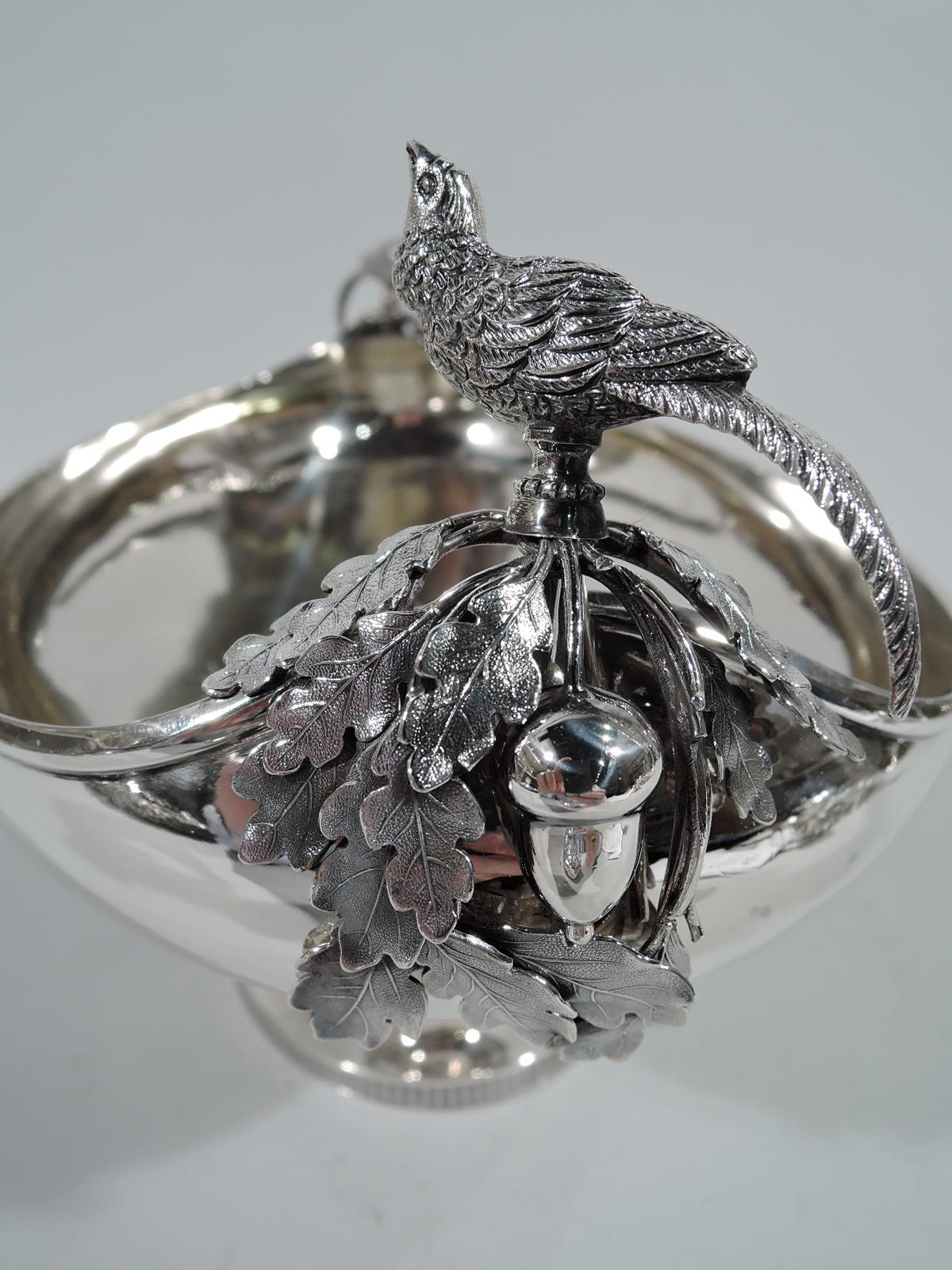 Aesthetic sterling silver centrepiece bowl. Oval with curved sides, short stem, and oval foot. On each end are applied oak leaves and acorn as well as perched bird with raised beak and warbling throat. Stylized ornament. Well centre has engraved