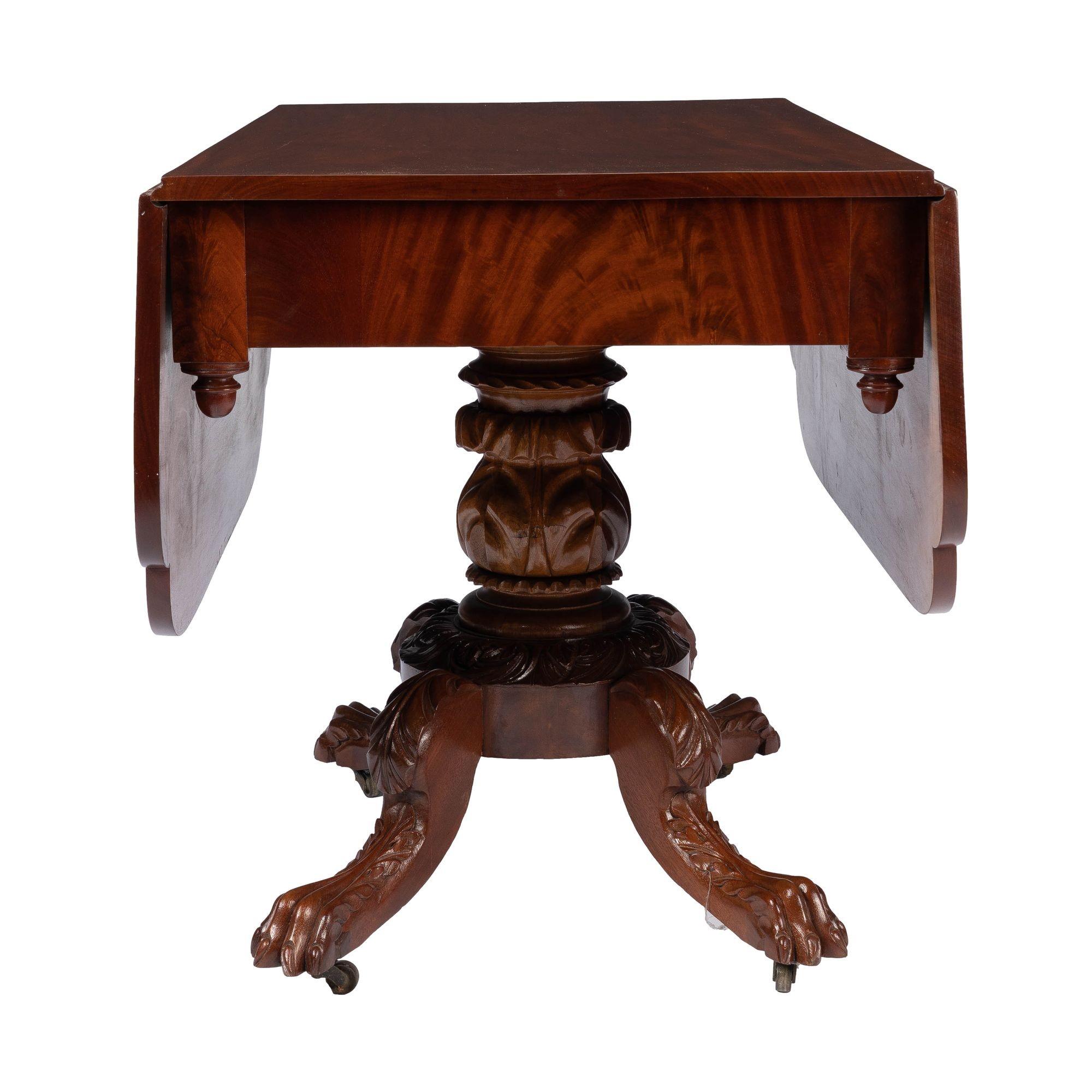American neoclassic drop leaf breakfast table with an acanthus carved pedestal base. This magnificent solid single board table top is mounted with two hinged drop leaves, cut with dimpled corners, and supported by pairs of butterfly brackets. The