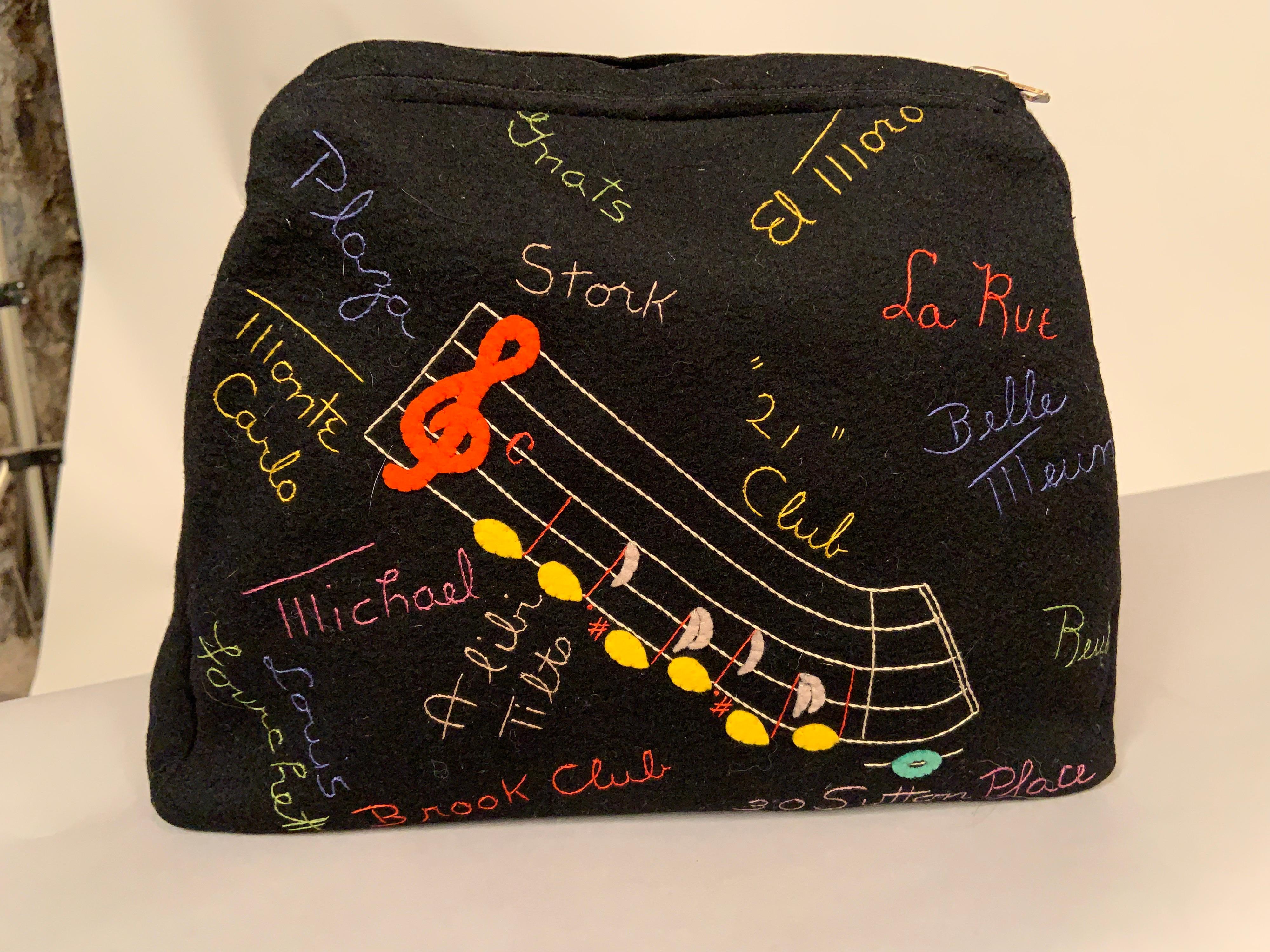A black wool felt clutch is appliqued on the front side with a cheerful yellow checker taxi cab carrying two passengers. It is surrounded by embroidered names of chic retail stores in  New York City, vacation destinations, race tracks etc. from the