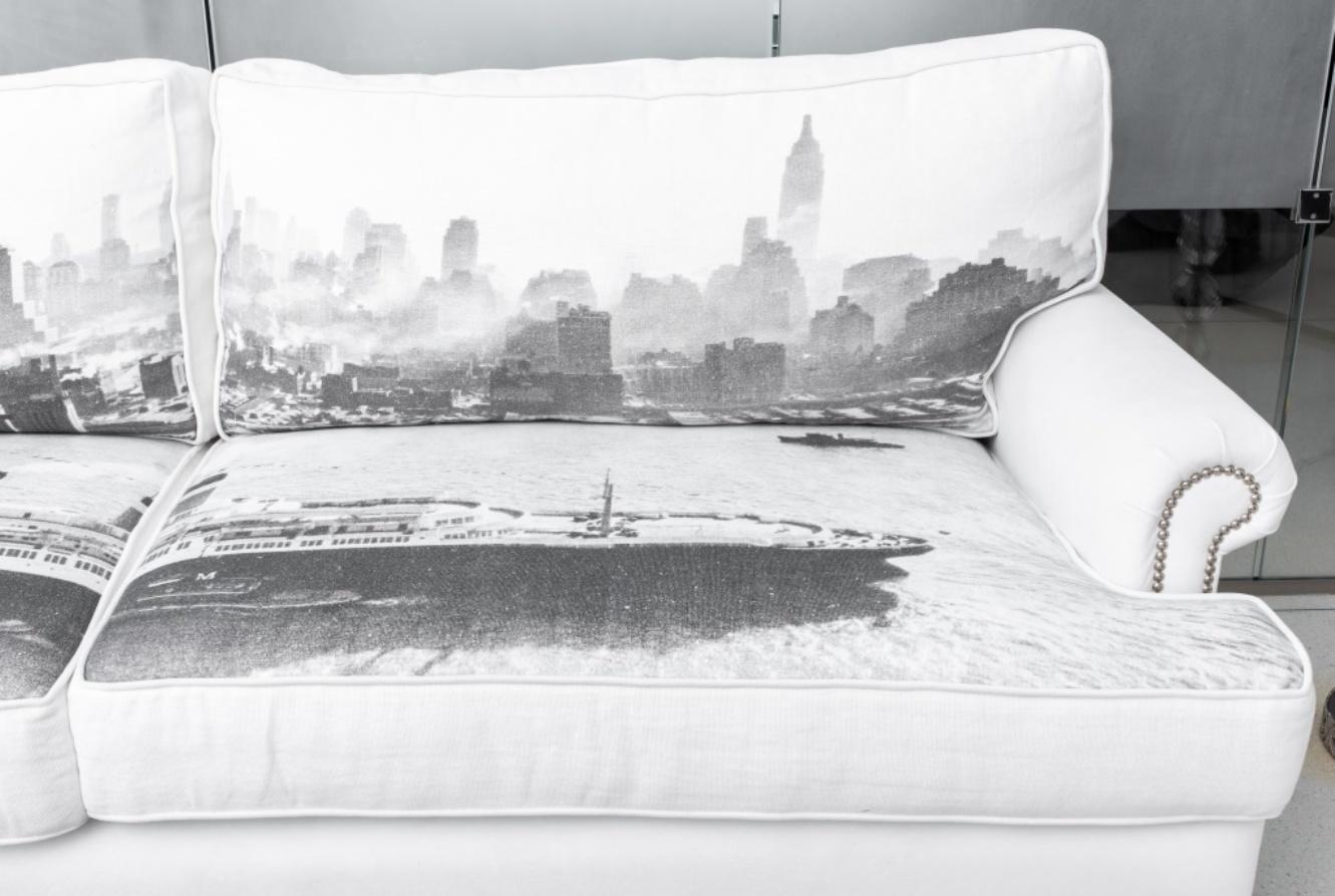 Three seat sofa upholstered in cream ultrasuede with canvas cushions printed with an early 20th century black and white photo of the skyline of New York City, studded with silver-tone studs, on four white legs. In good condition. Wear consistent