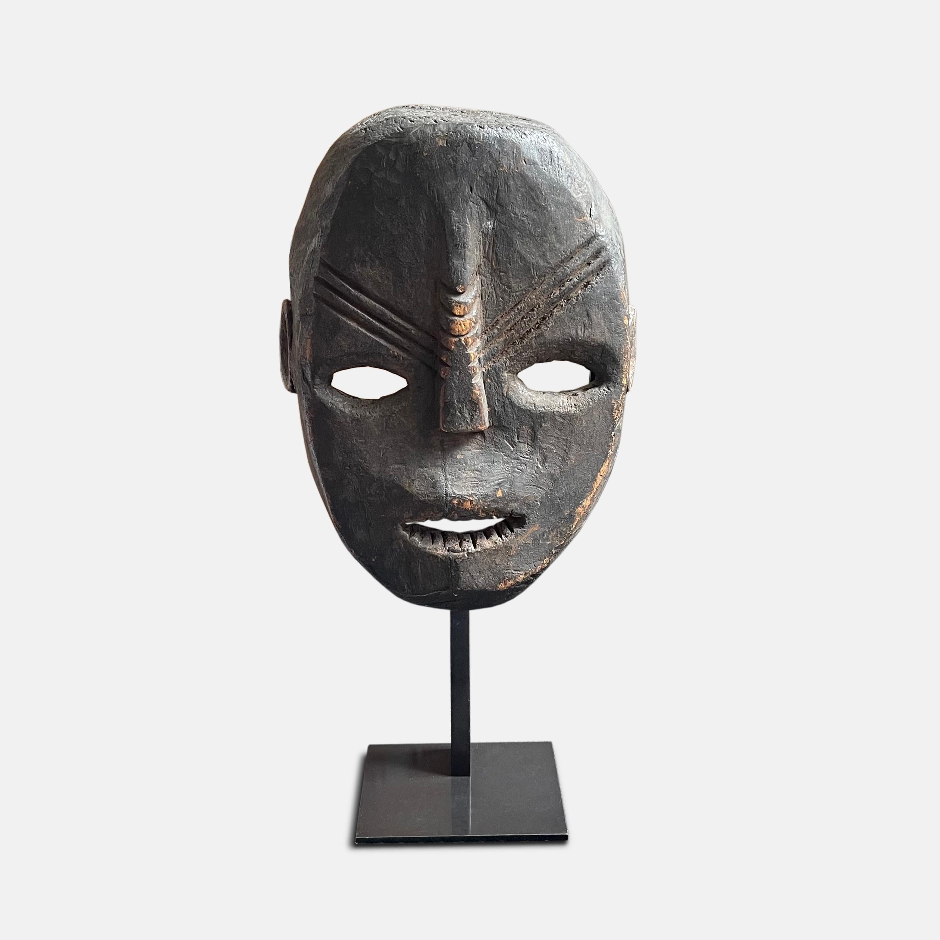 With a beautiful dark patina, this early 20th century wooden mask shows a naturalistic human face, flattened, with the forehead, open mouth and stylised teeth in light relief; the ridged, elongated nose and the diagonal ridges on the forehead are