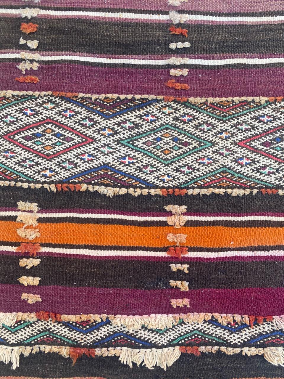 Entdecken Sie die zeitlose Eleganz eines marokkanischen Berber-Kilims aus dem frühen 20. Jahrhundert. Dieser exquisite flache Teppich besticht durch sein geometrisches Stammesmuster in atemberaubenden Naturfarben, darunter Lila, Orange, Grün, Blau,
