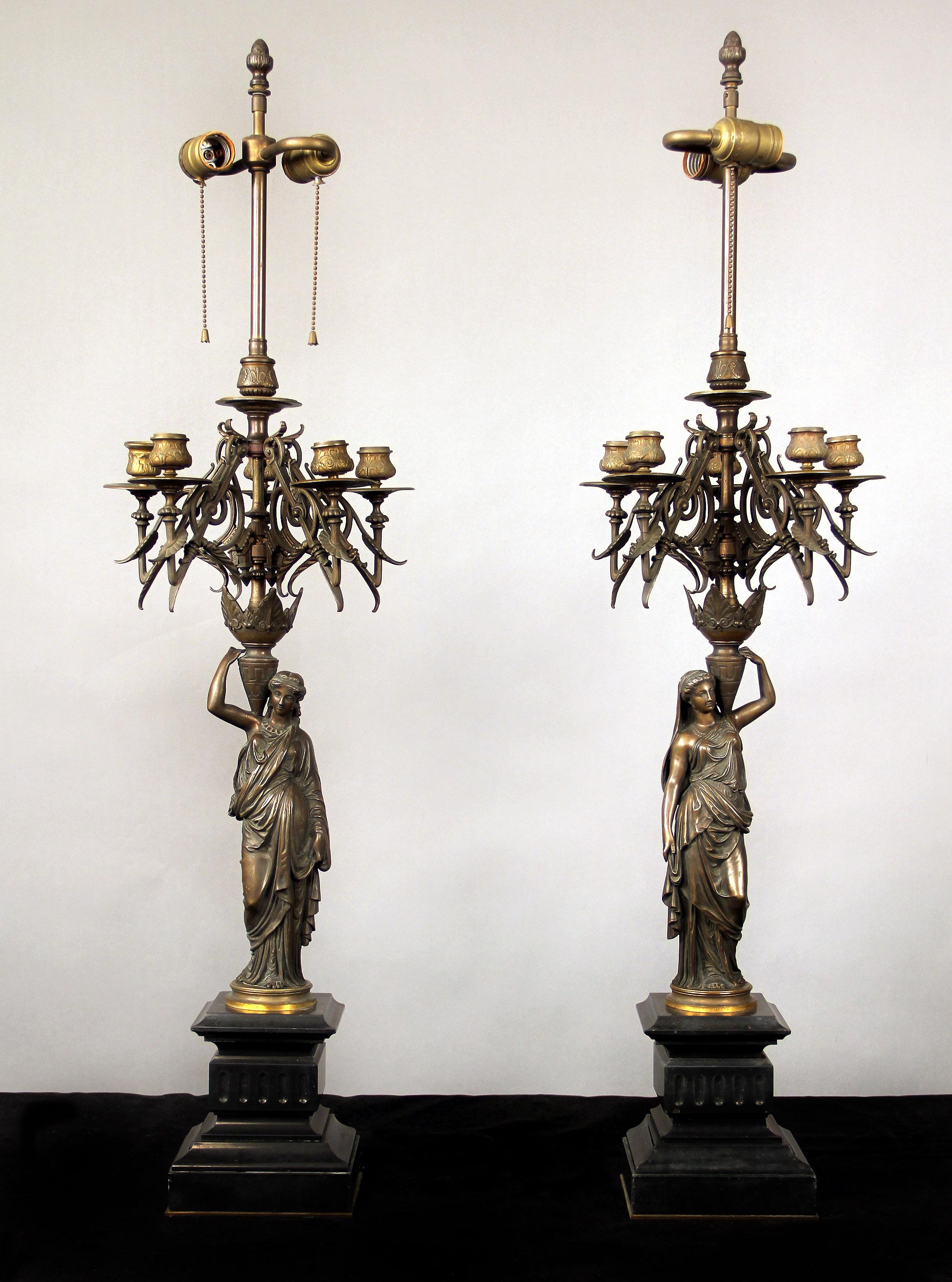 A nice pair of late 19th century patina bronze two light figural candelabra lamps

Each cast as a female figure in draped garment upholding candelabra with five perimeter arms and one centered leading up to two lights, standing on a black marble