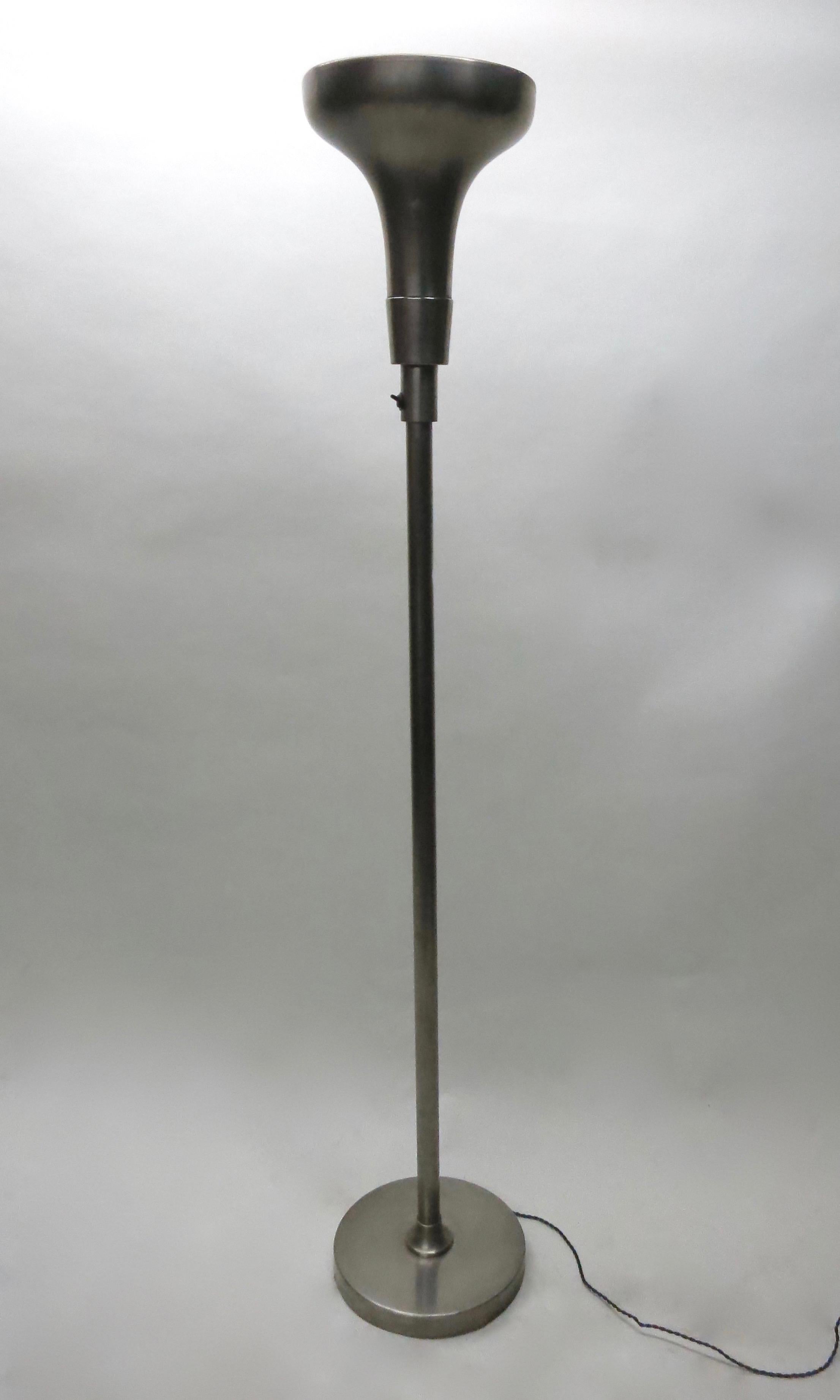 Deco floor lamp in nickel-plated copper and steel with a round base, and a tubular stem that connects to the torchiere shade that encompasses a single light socket. The black flip switch is located below the bottom portion of the shade's perforated