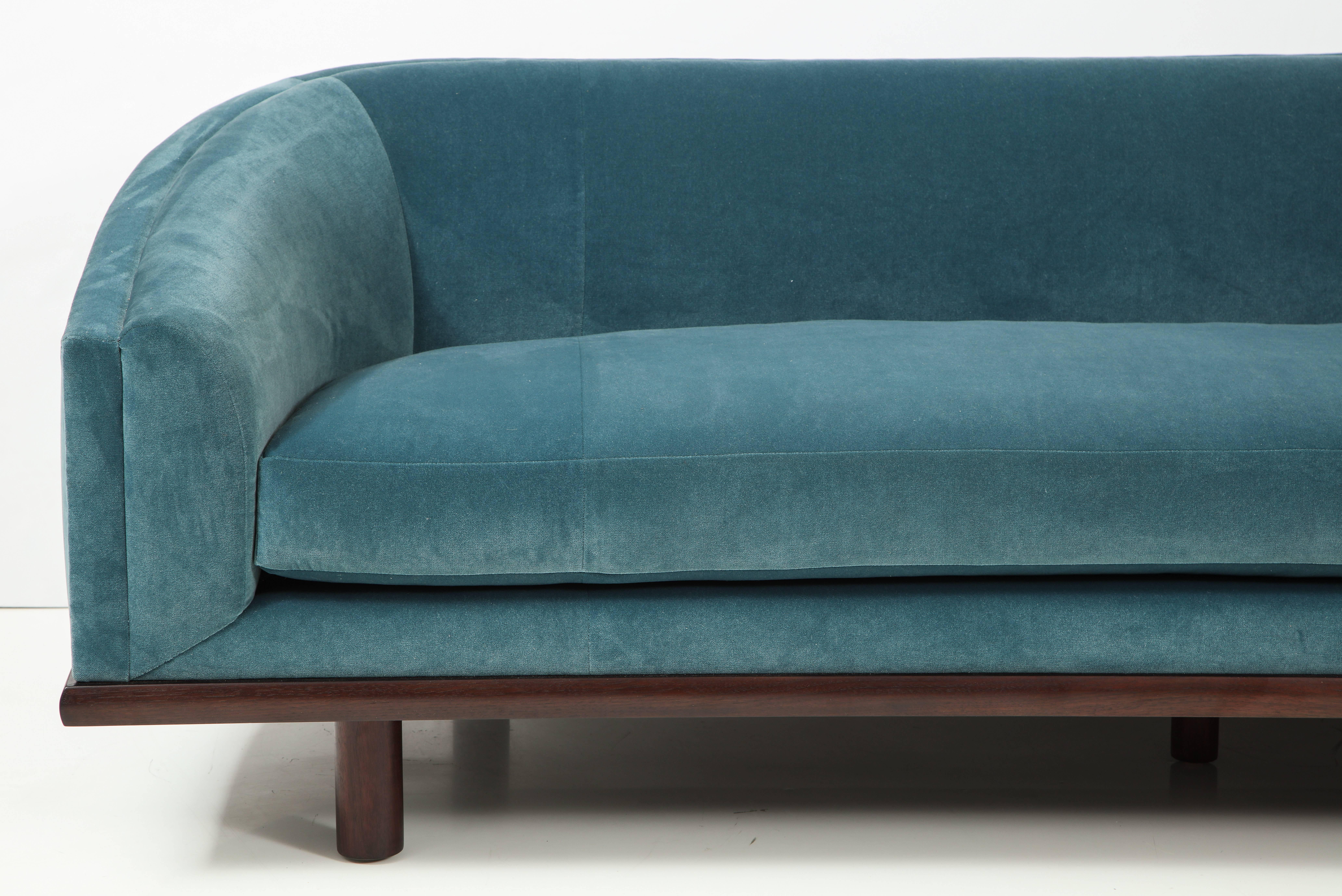 Elegant tight back sofa with curved silhouette and long single seat cushion on solid wood framed base and Classic minimal cylinder legs. The sofa is upholstered in a teal blue velvet with a mahogany base.
