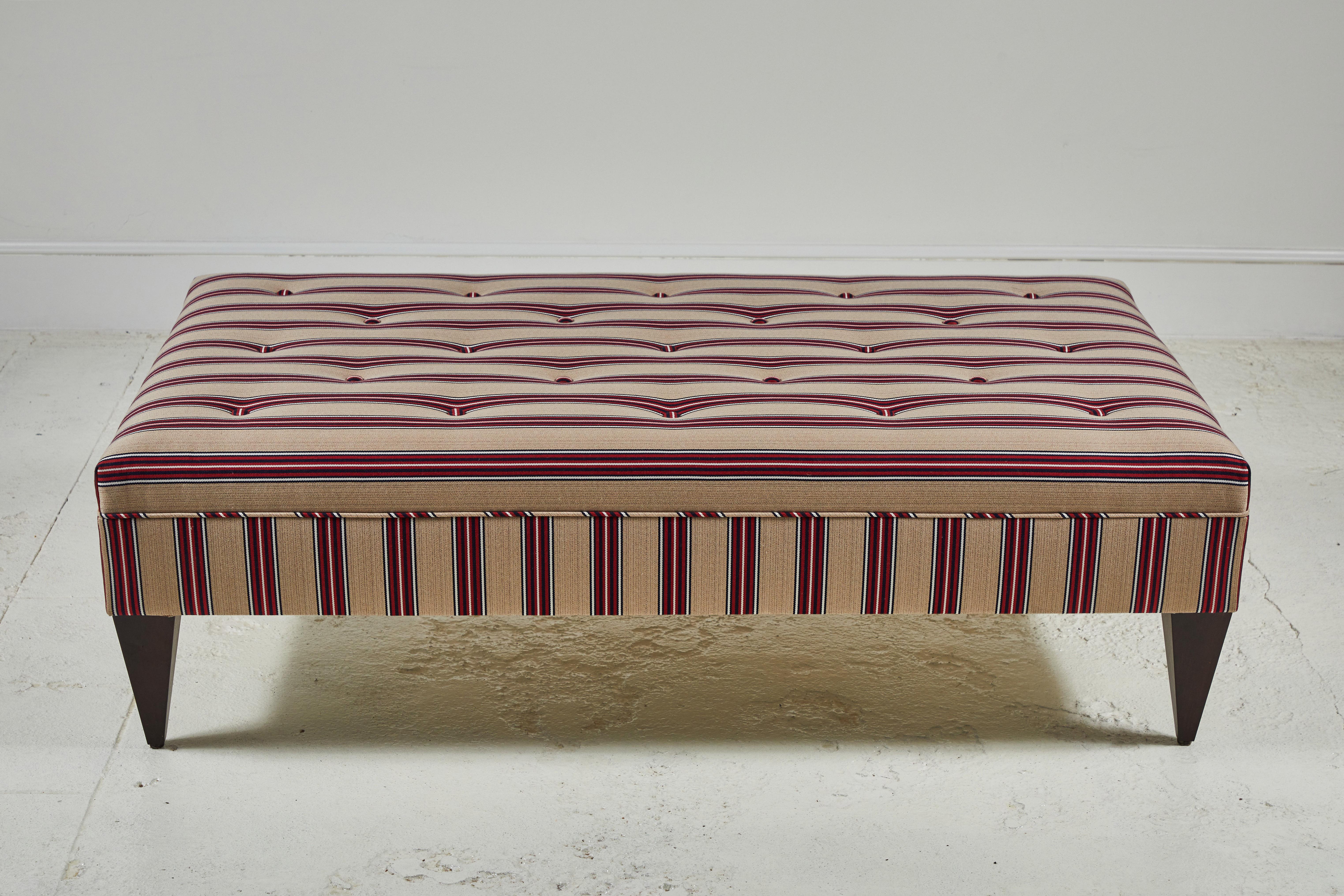 Simple and straight forward ottoman with tufting along the top, and one row of piping. Finished off with tapered legs. This specific ottoman is upholstered in striped fabric from UK textile company, Howe.
