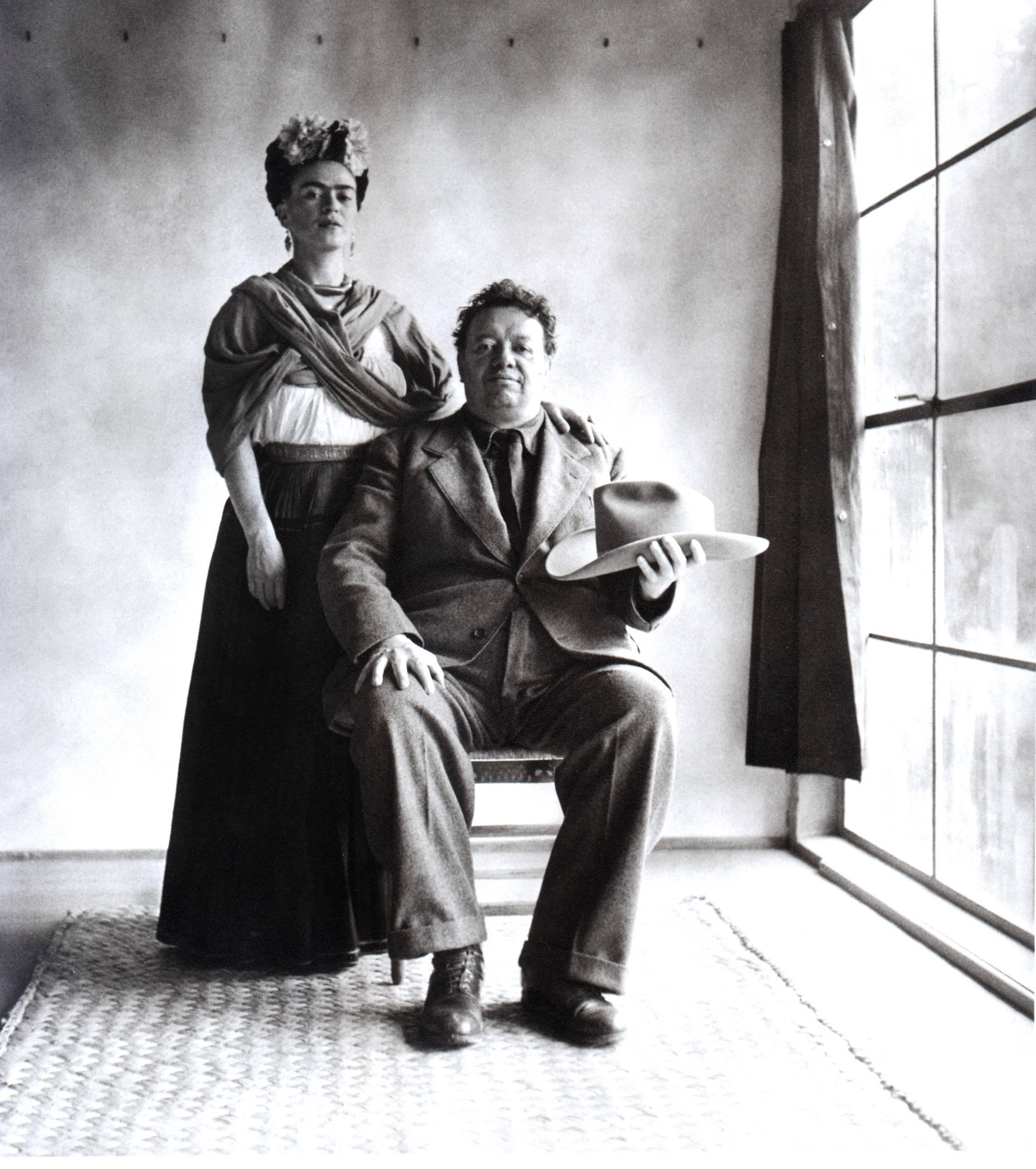 Frida et Diego avec chapeau de Nickolas Muray représente un couple posant pour un portrait. Frida Kahlo est debout à côté de Diego Rivera, qui est assis et tient un chapeau à la main. Ils se trouvent dans une pièce vide, avec une grande fenêtre