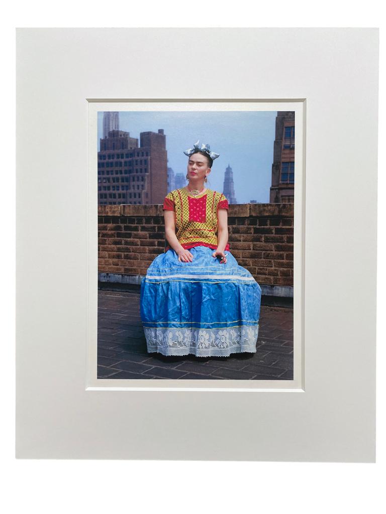 Frida in New York by Nickolas Muray features Frida Kahlo in a colorful red, yellow and blue dress with light blue bows in her hair and a cigarette in her hand. She is seated on top of a building, with part of the New York skyline visible in the