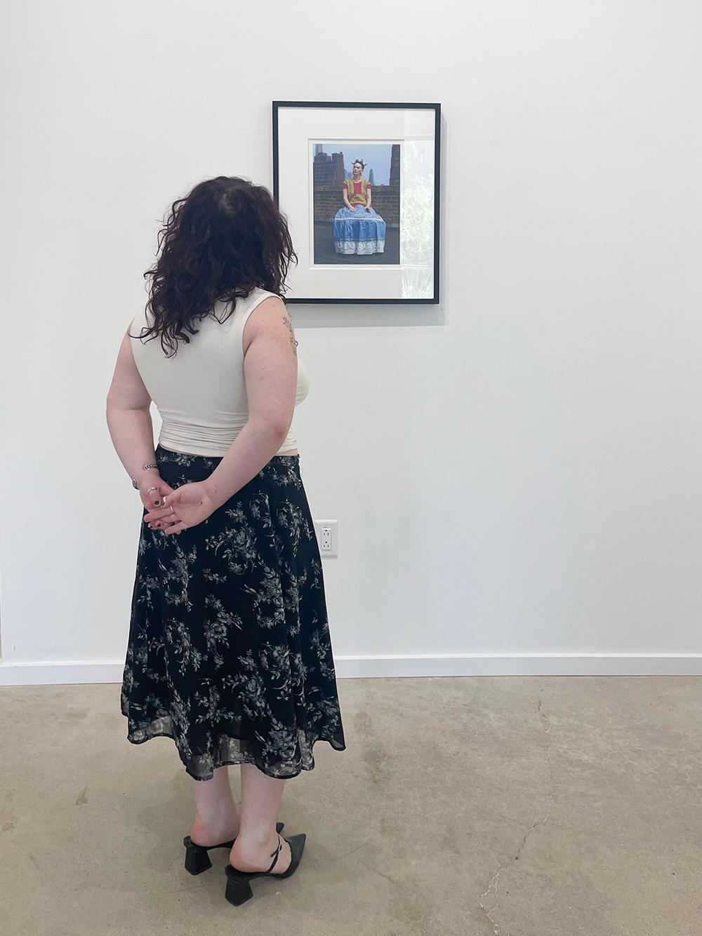 Frida in New York von Nickolas Muray zeigt Frida Kahlo in einem farbenfrohen roten, gelben und blauen Kleid mit hellblauen Schleifen im Haar und einer Zigarette in der Hand. Sie sitzt auf dem Dach eines Gebäudes, im Hintergrund ist ein Teil der New