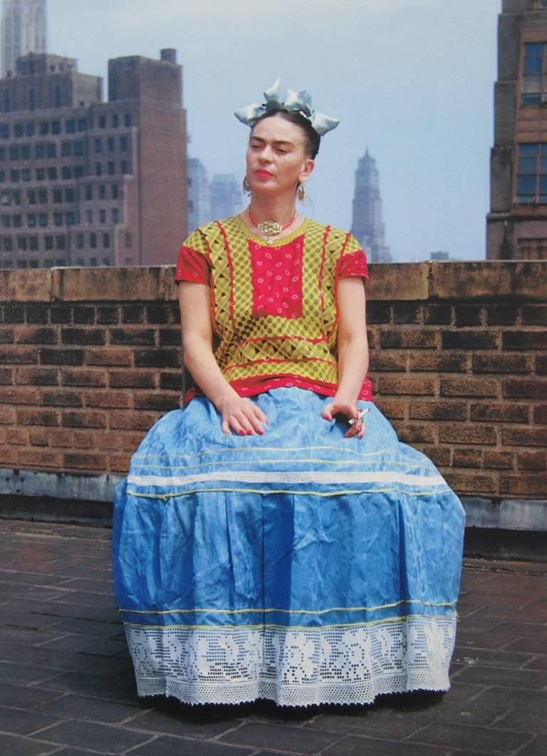 Frida in New York by Nickolas Muray features Frida Kahlo in a colorful red, yellow and blue dress with light blue bows in her hair and a cigarette in her hand. She is seated on top of a building, with part of the New York skyline visible in the