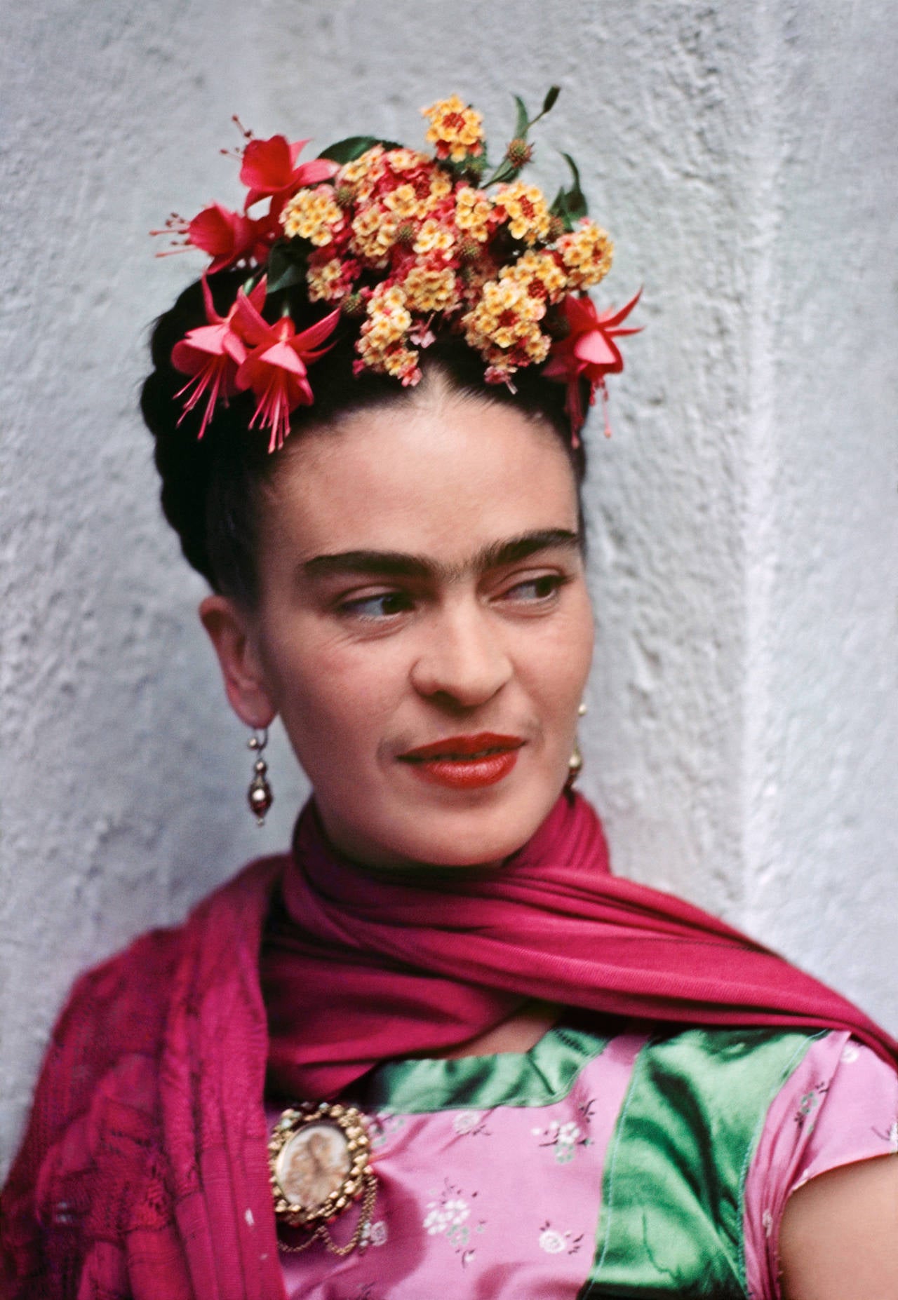 Frida in Pink and Green Blouse by Nickolas Muray is a colorful portrait of Mexican painter Frida Kahlo. Frida wears a green and pink shirt with a magenta scarf wrapped around her neck. She wears yellow and pink flowers in her hair, and glances off
