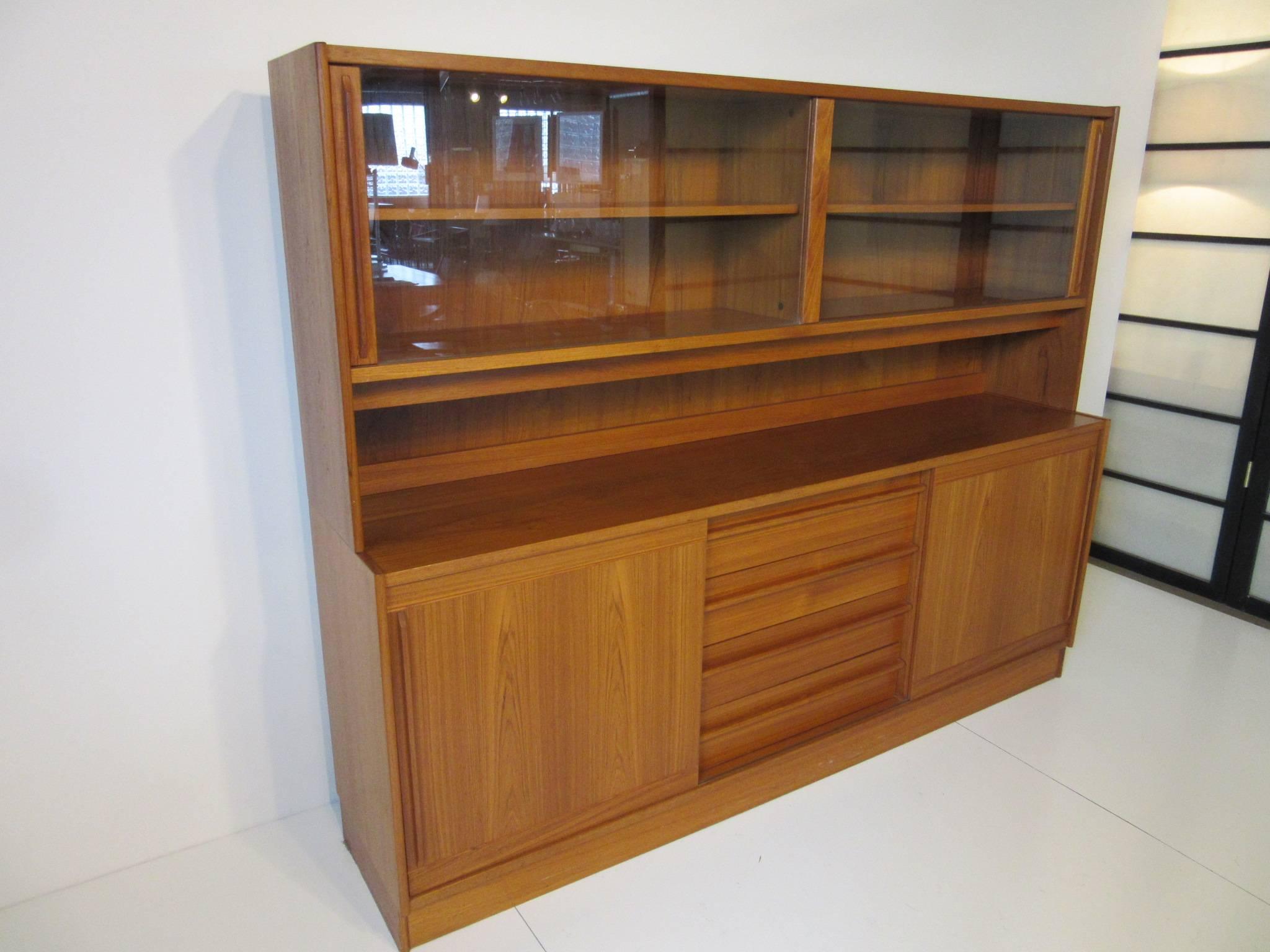 A Danish teak wood two-piece cabinet with upper display or bookcase top, two sliding glass doors with matching trim and a credenza cabinet below with four drawers to the front and sliding doors to each side. Inside storage with two adjustable