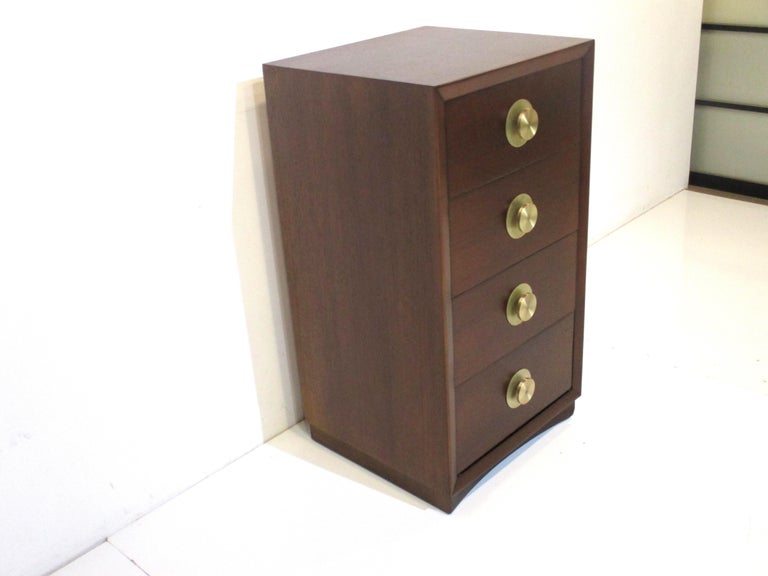 A taller nightstand or thin small chest having four drawers the top one with dividers and a small moveable tray . The turned brass pulls and backing plates and dark walnut toned wood give the piece a rich and stylish feel , manufactured in the