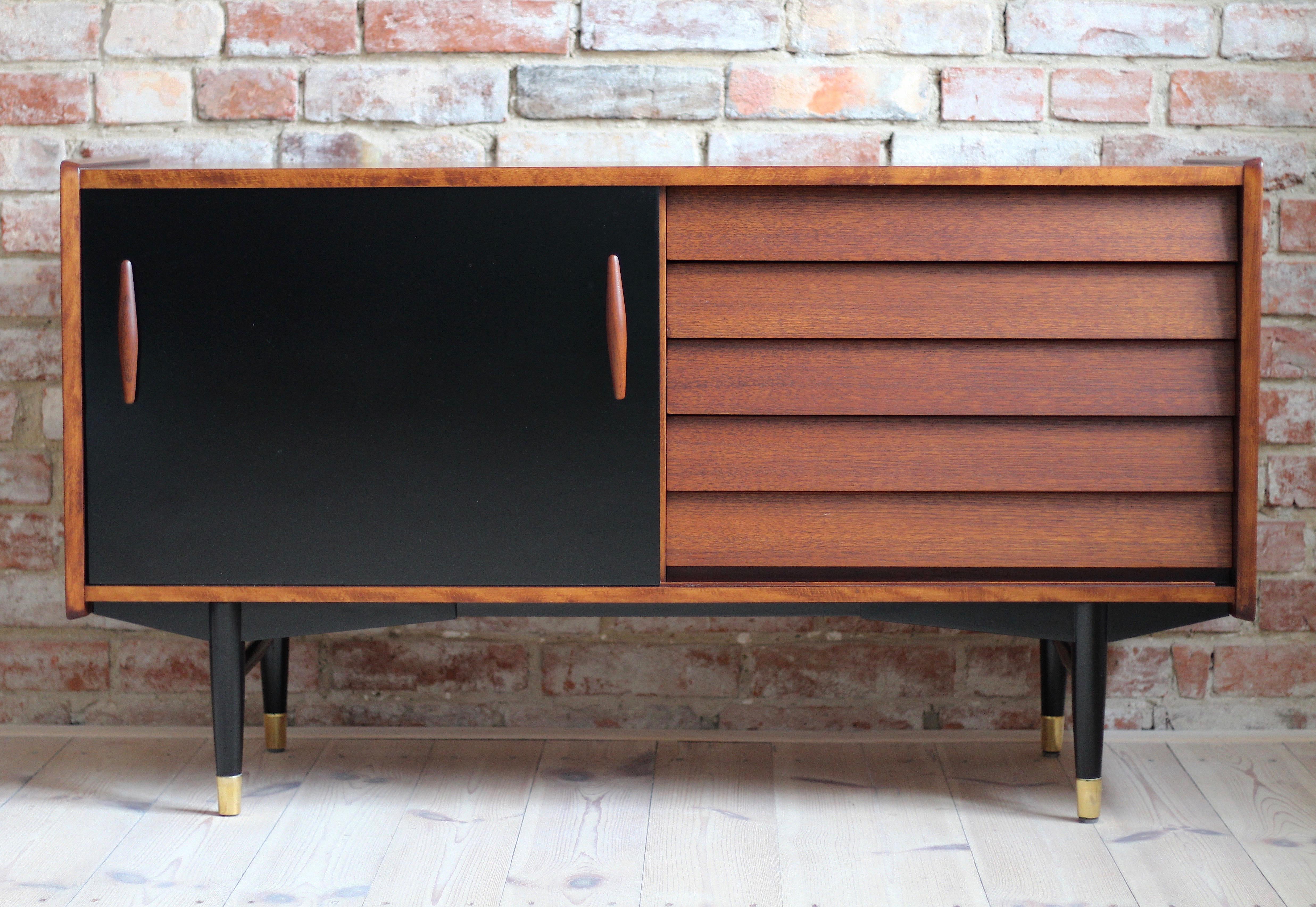 This is a not so common model of sideboard by Nils Jonsson in teak wood with black sliding door, two sculpted handles, elegant black base completed with subtle brass legs cups - a great example of Scandinavian Modern style in its best. Easily