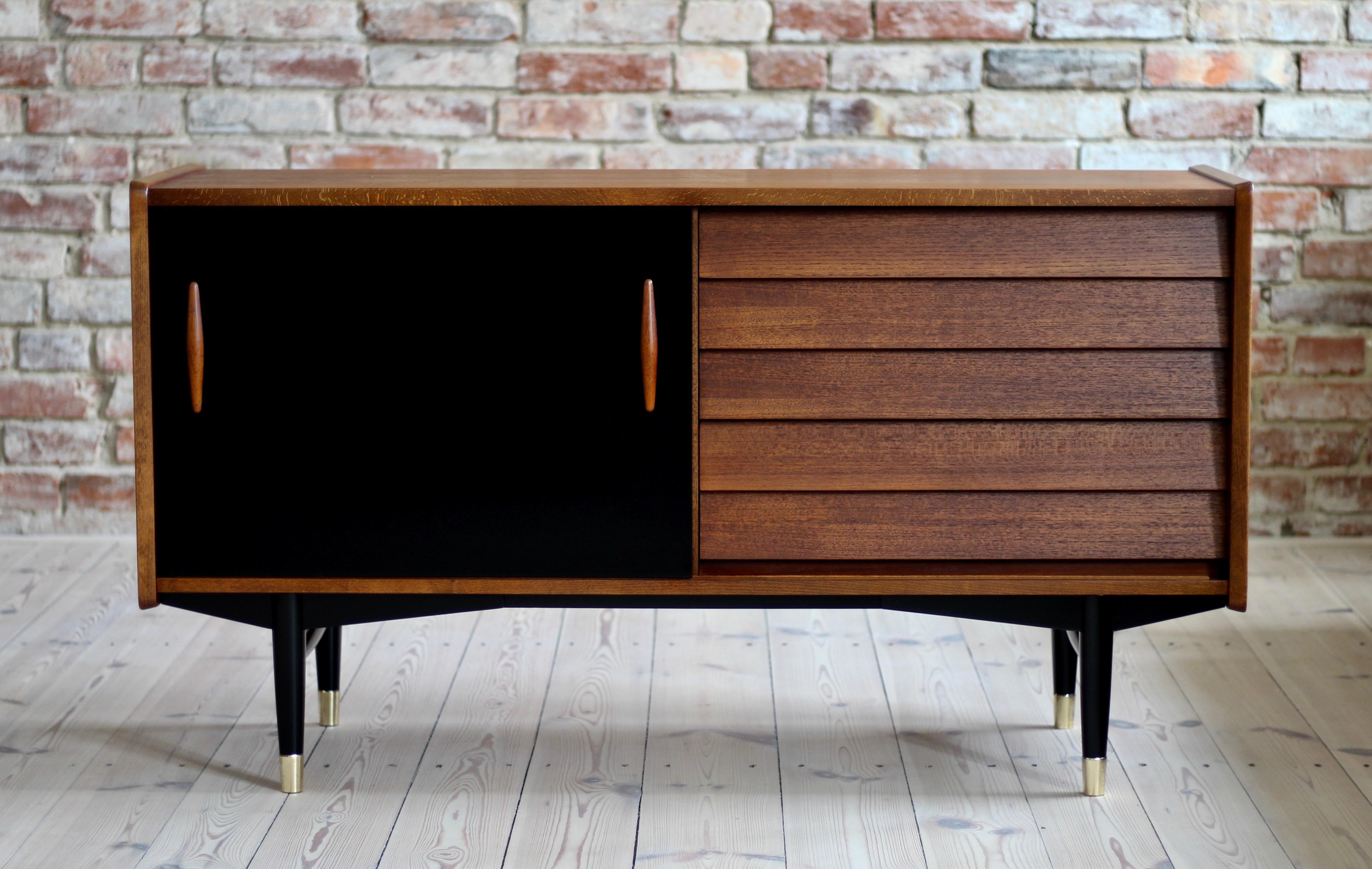 This is a not so common model of sideboard by Nils Jonsson in teak wood with black sliding door, two sculpted handles, elegant black base completed with subtle brass legs cups - a great example of Scandinavian Modern style in its best. The piece was