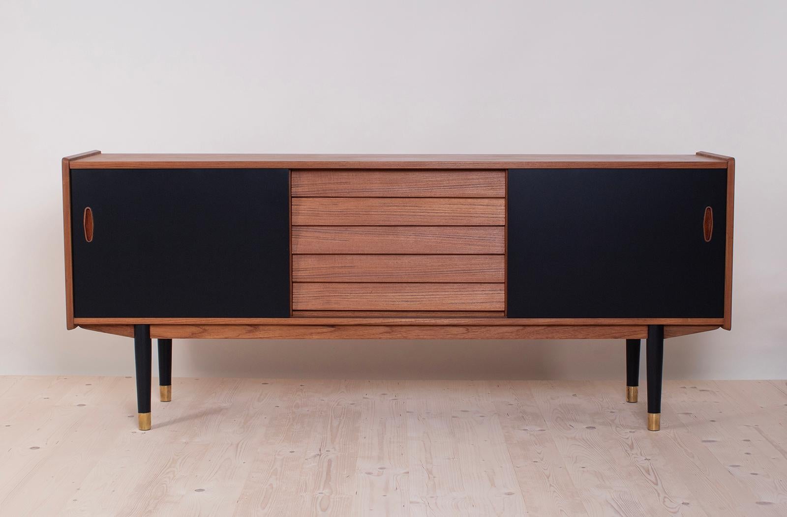 This is not so common model of sideboard by Nils Jonsson in teakwood with black sliding door, two sculpted handles, elegant base completed with subtle slender legs, all 4 legs are finished with brass – a great example of Scandinavian Modern style in