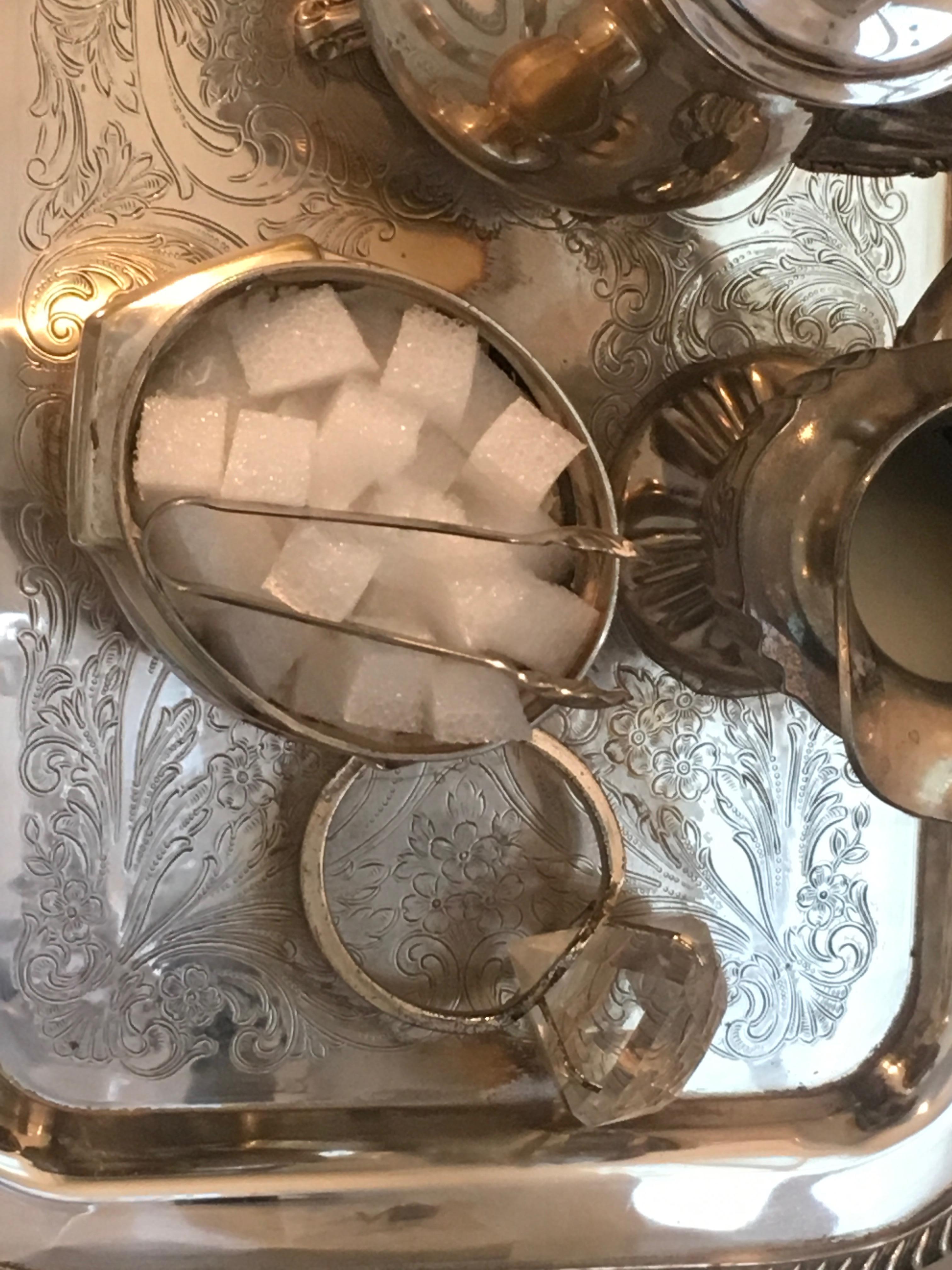 Entitled VI by Nina Bentley is a mixed media sculpture created with a  silver metal tray, metal teapots, coffee pots, a sugar server and milk server plus resin and styrofoam. There is also a large ring on the tray.   It is an original, one of a kind