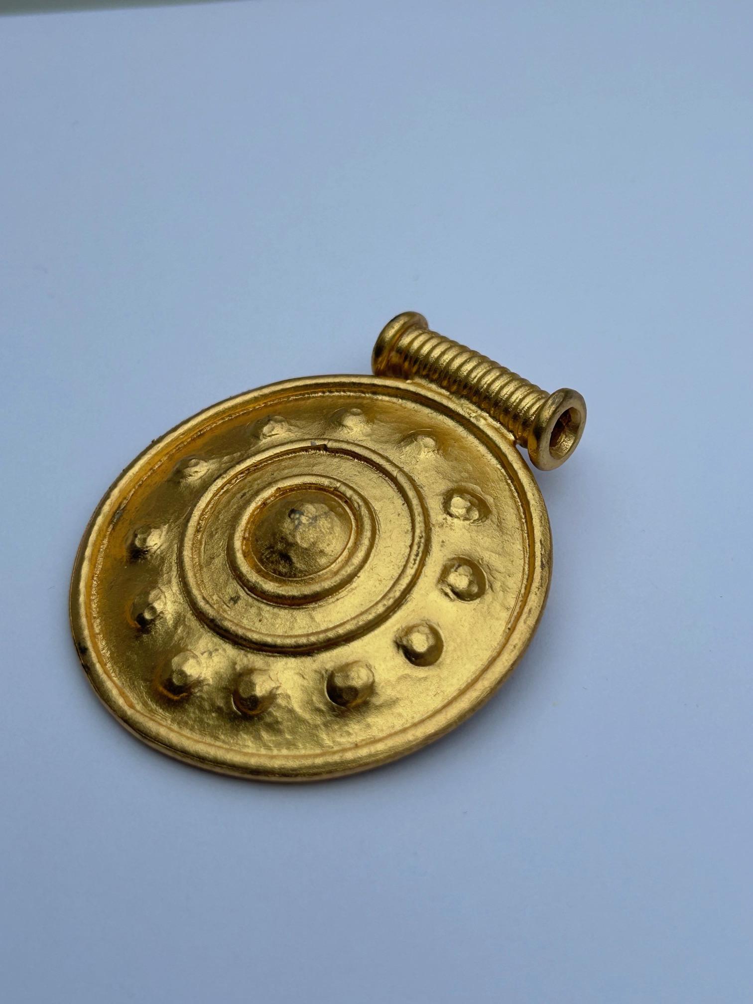 Nuestro broche estilo botón de Nina Ricci, vintage y muy atractivo, en metal dorado, también se puede llevar como collar si se pasa una cadena (no incluida) por la parte superior. Con unas medidas de 52 mm a lo ancho del círculo y 59 mm incluida la