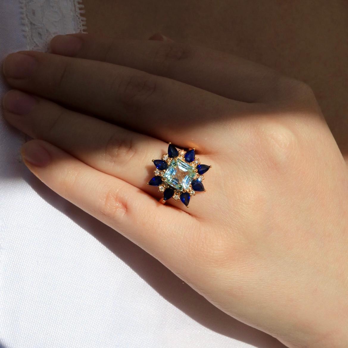 Unveil a masterpiece of elegance with the Margaret Aquamarine Sapphire Diamond Cocktail Ring. At its heart lies a princess-cut aquamarine, exuding tranquil oceanic hues and unmatched brilliance. Encircling this captivating gem, pear-cut royal blue