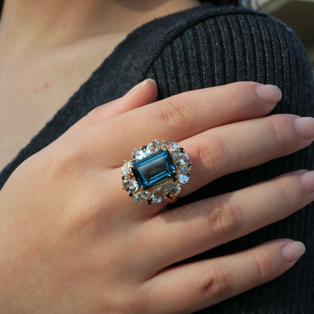 Ti presentiamo l'anello da cocktail Margaret London Blue Topaz Aquamarine Sapphire, un capolavoro di eleganza e opulenza. Realizzato in lussuoso oro massiccio, questo anello ha al centro uno splendido Topazio Blu di Londra dal taglio a smeraldo, che