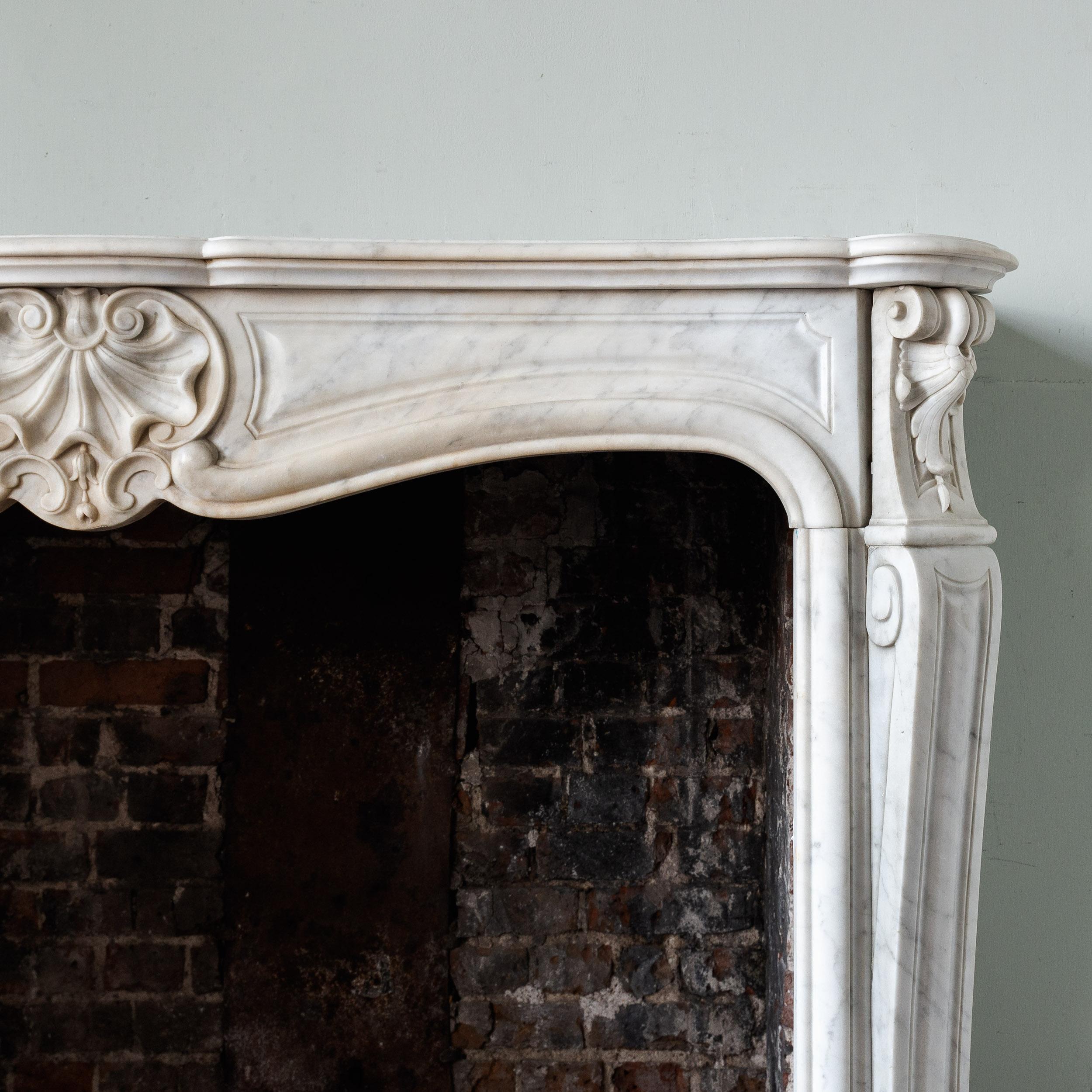 A good late 19th century French Louis XV style fireplace in lightly veined Italian Carrara marble, the deep, shaped and moulded shelf above frieze centred by elegantly carved scallop shell, the canted volute jambs headed by foliate clasps, on block
