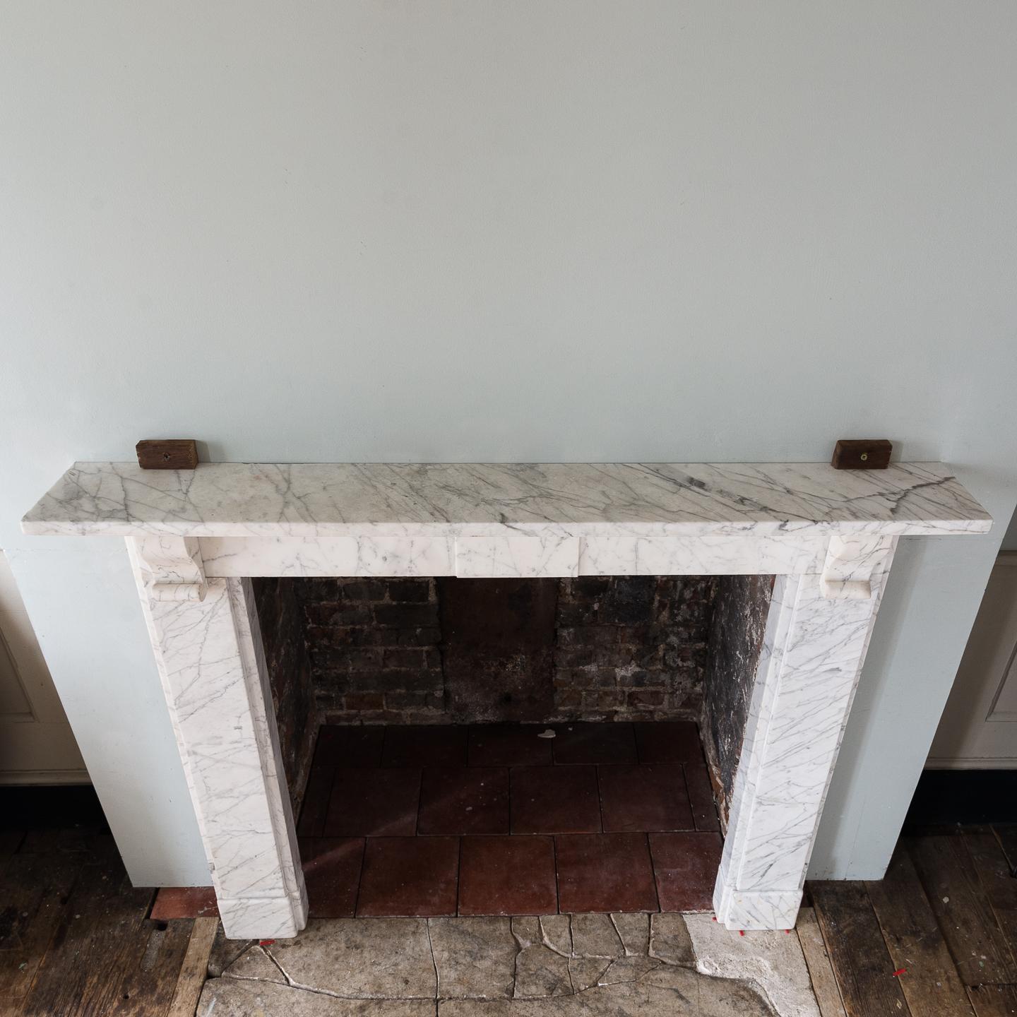 19th century ‘Old English’ marble chimneypiece, the rectangular shelf above over plain frieze with plain central tablet, the jambs headed by simple corbels, on footblocks.
   
  