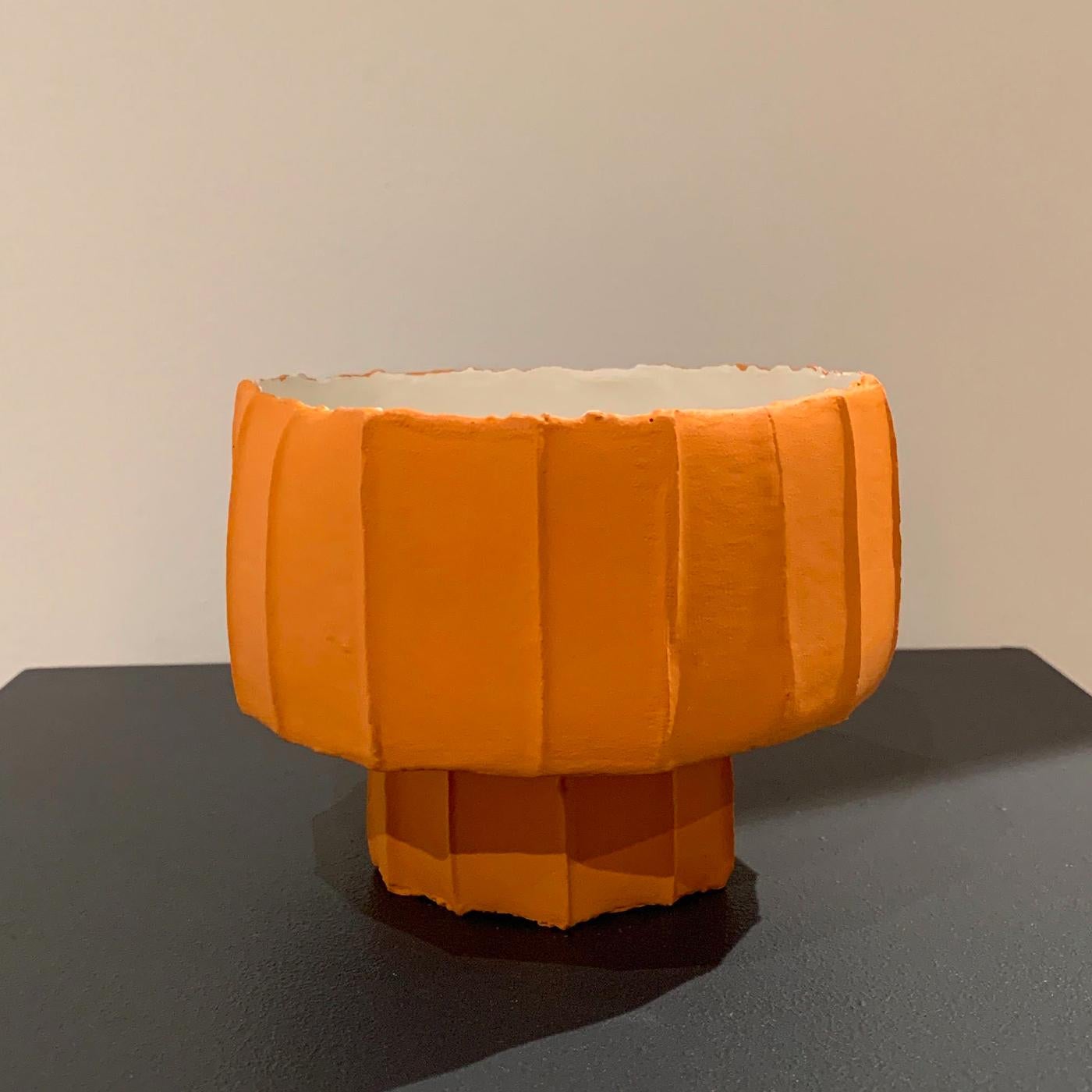 Marked by a vibrant light orange color, this vase has a distinctive silhouette that defies classical artistic norms: composed of two, inverted cylindrical tiers, the vase has a large tier stacked atop a small, narrow base. The vase is handmade using