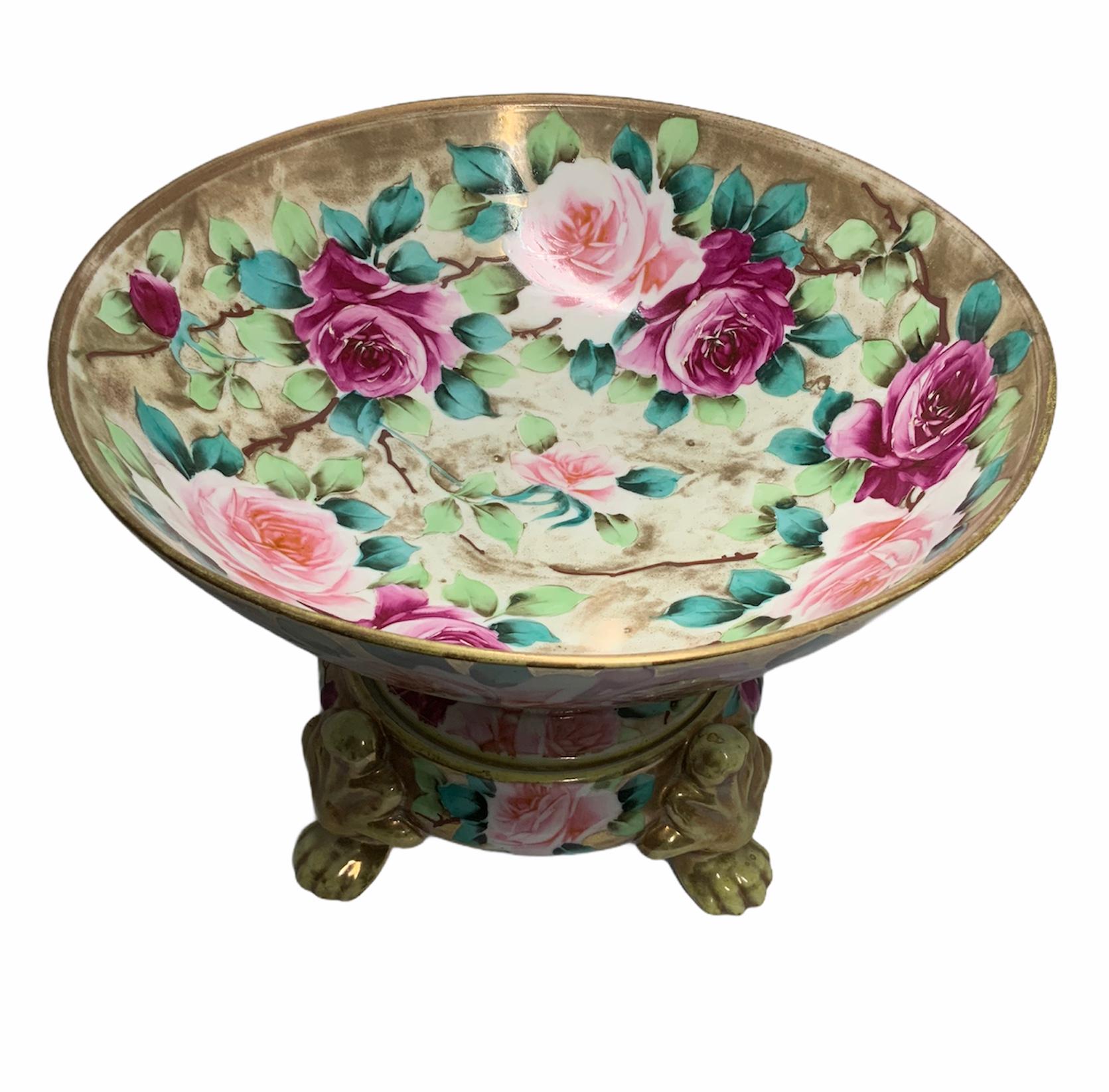 This a Nippon porcelain wide and round gilt bowl hand painted with large pale & dark pink roses with green foliage. The bowl stands in a round base with four lion’s paws. Under the base is the Nippon hallmark and is written “hand painted”.