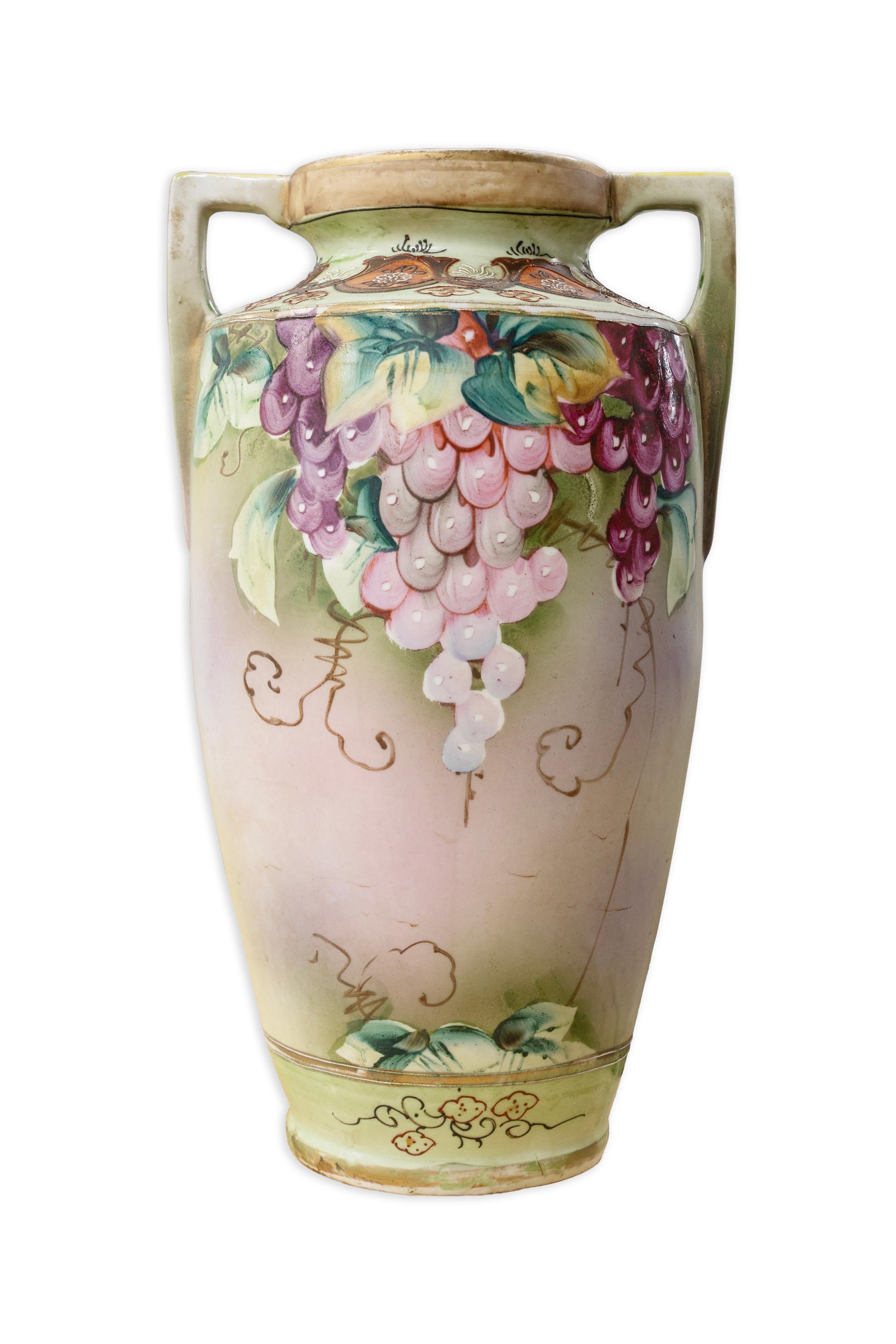 A charming pair of early 20th century hand painted Nippon porcelain vases, each featuring twin handles and a richly decorated design of cascading grape clusters in shades of purple, rose, and white, accented with vibrant green foliage. The bodies