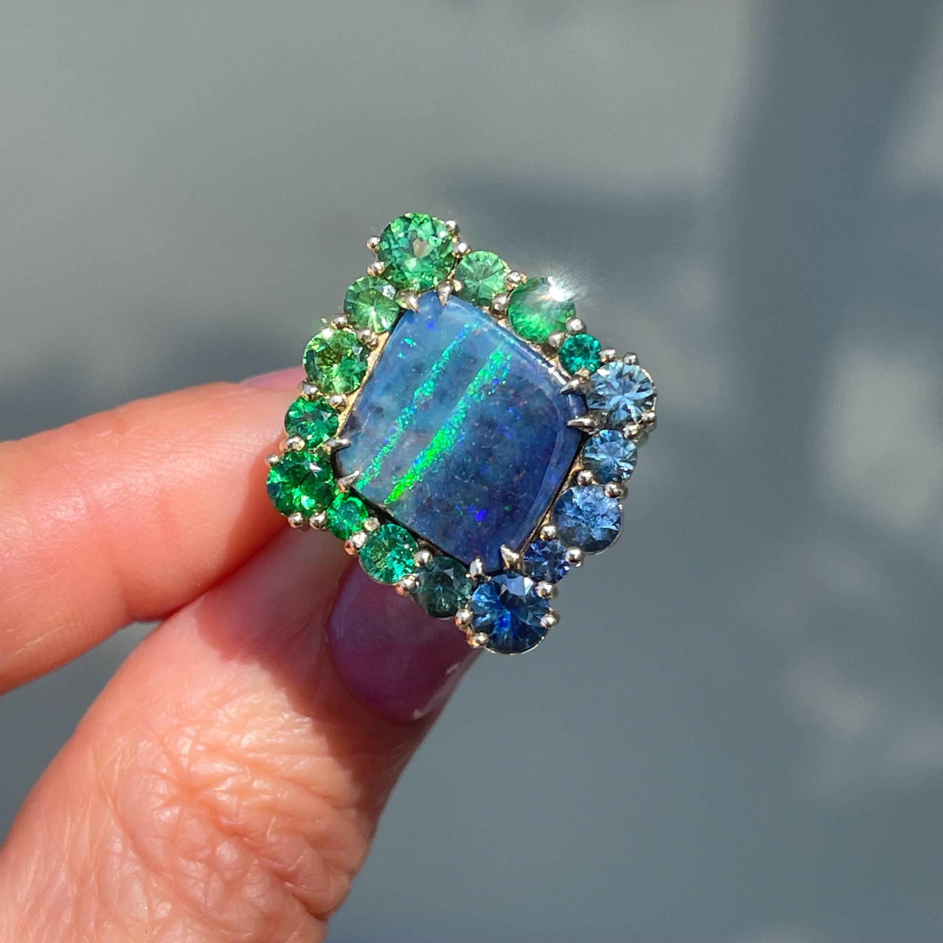 Verdant greens traverse the steely blue rhomboid that shapes this Australian Opal Ring. Framed by a halo of sapphires, emeralds and tsavorite garnets, the Boulder Opal sees its palette reflected in the surrounding stones. Held in place by golden