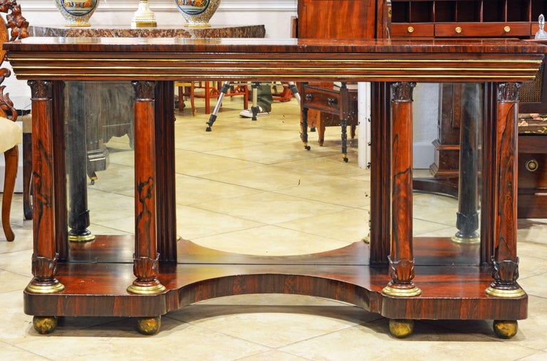 Great classic design and the beauty of the deep colored rosewood makes this console table both stately and refined. The polished top rests on a brass trimmed molded frieze supported by round corinthian style columns with archantus carved bases on