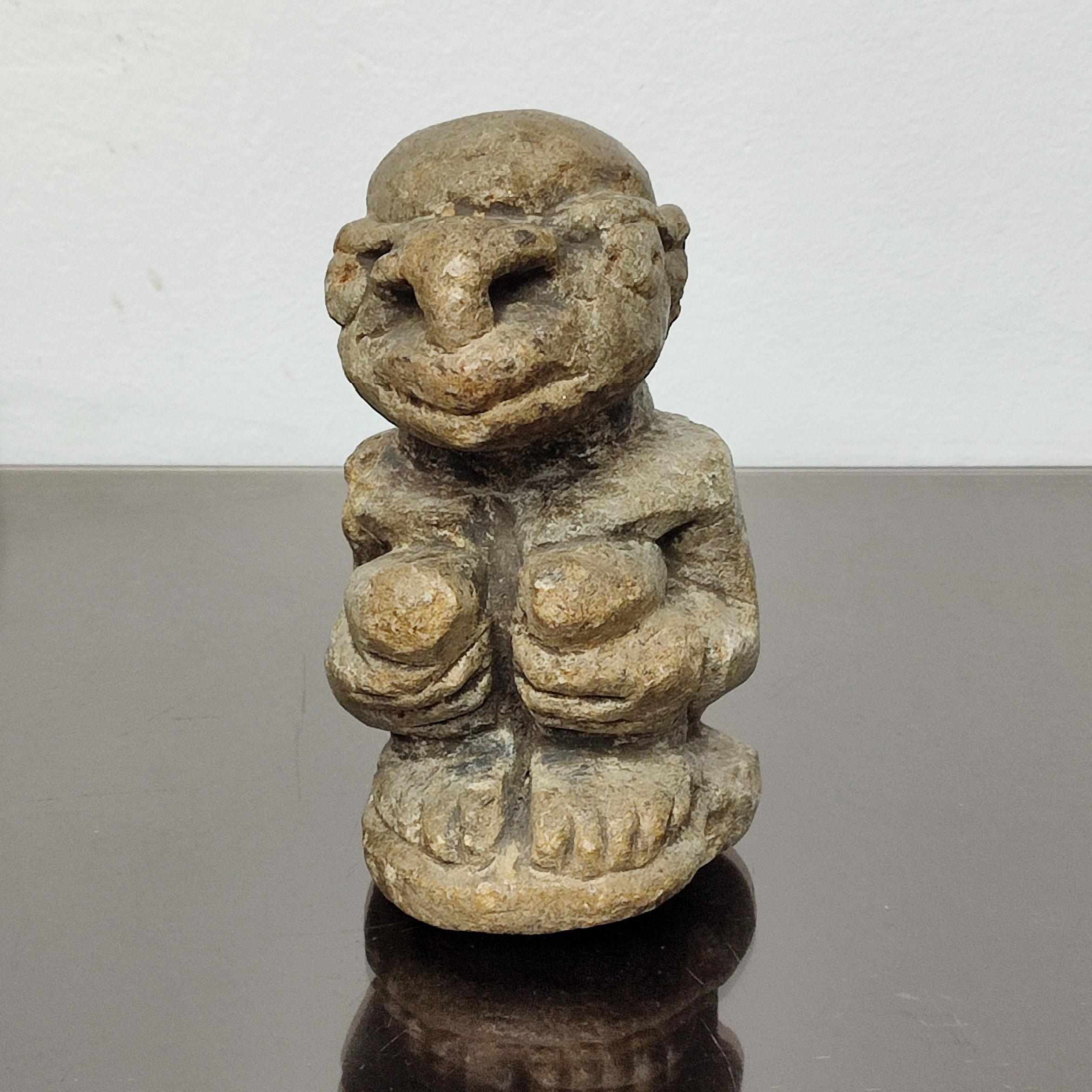 Eine Kissi Nomoli-Steinfigur, ein Mann mit um die Knie geschlungenen Händen, auf einem Sockel; vom Volk der Kissi in Sierra Leone.
Graubraune Patina, schnauzenartiger Mund, große Nase, leichte Abriebspuren.
Solche Figuren wurden in