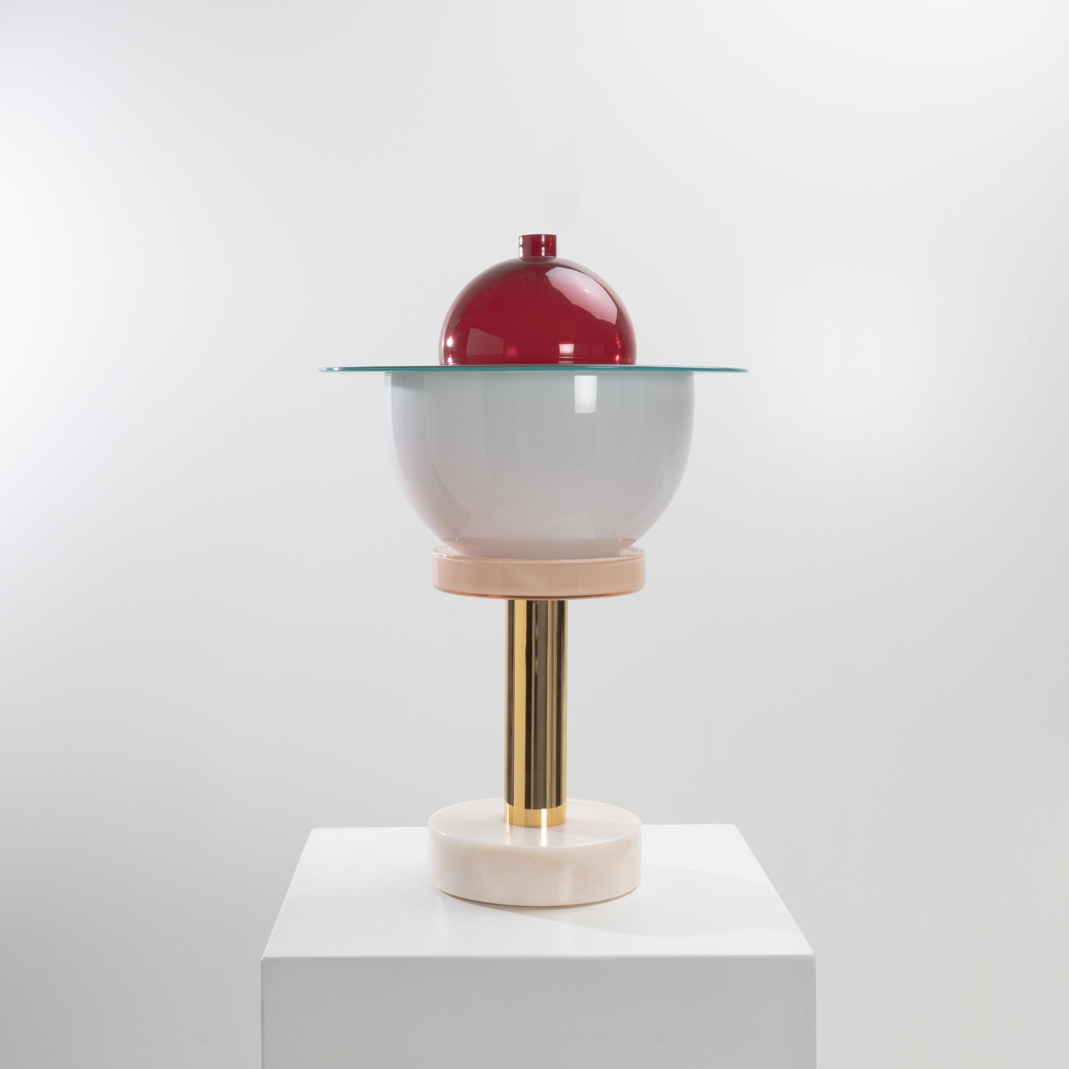 Designed in 1994 by the great architect-designer Ettore Sotssass, Nopuram is a light composed of a pink marble base surmounted by a golden steel shaft.
4 blown glass elements surmount the whole, a pink glass base, a milk white glass cup which