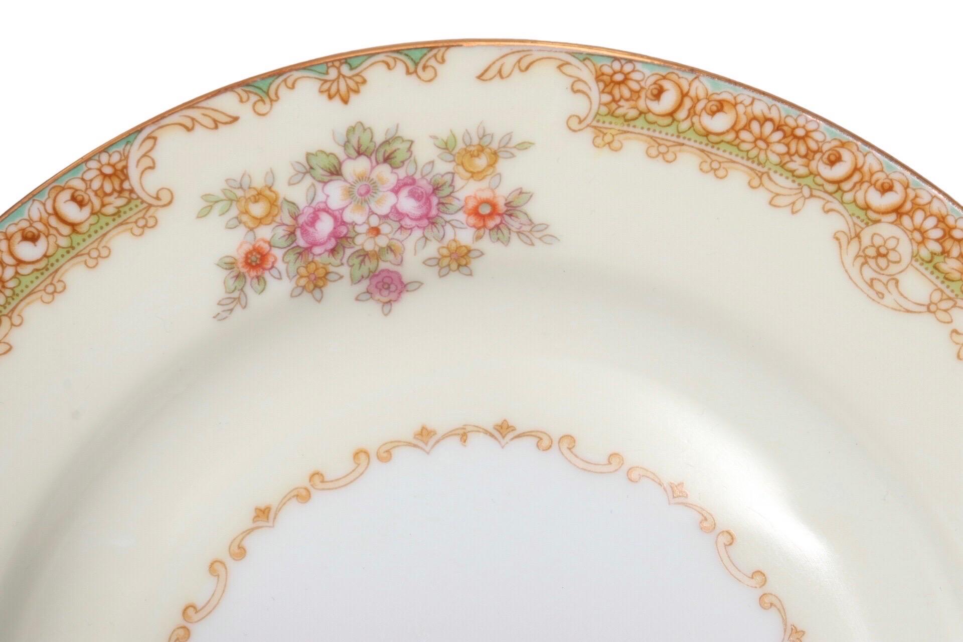 A set of twelve Japanese dinner and side plates made by Noritake in their N134 pattern, which was discontinued circa 1940. An elaborate border is hand painted with peonies, serpentine scrolls with acanthus trim and interspersed with colorful floral