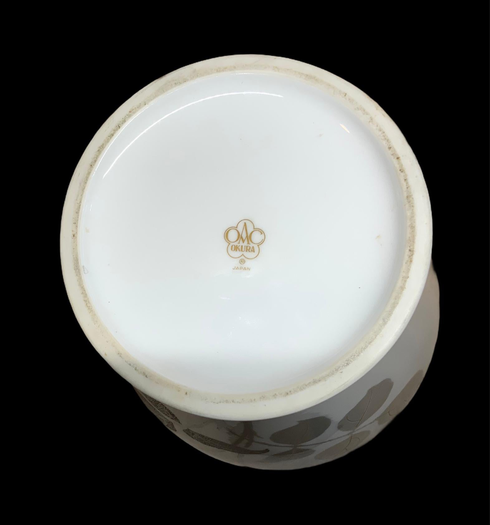 This a Noritake, Okura porcelain flower vase hand painted white in the background with a large gold and silver etching rose in the front and rose bud branches in the back. Also the rim is painted gold. Under the base is hallmarked Okura, Japan (one
