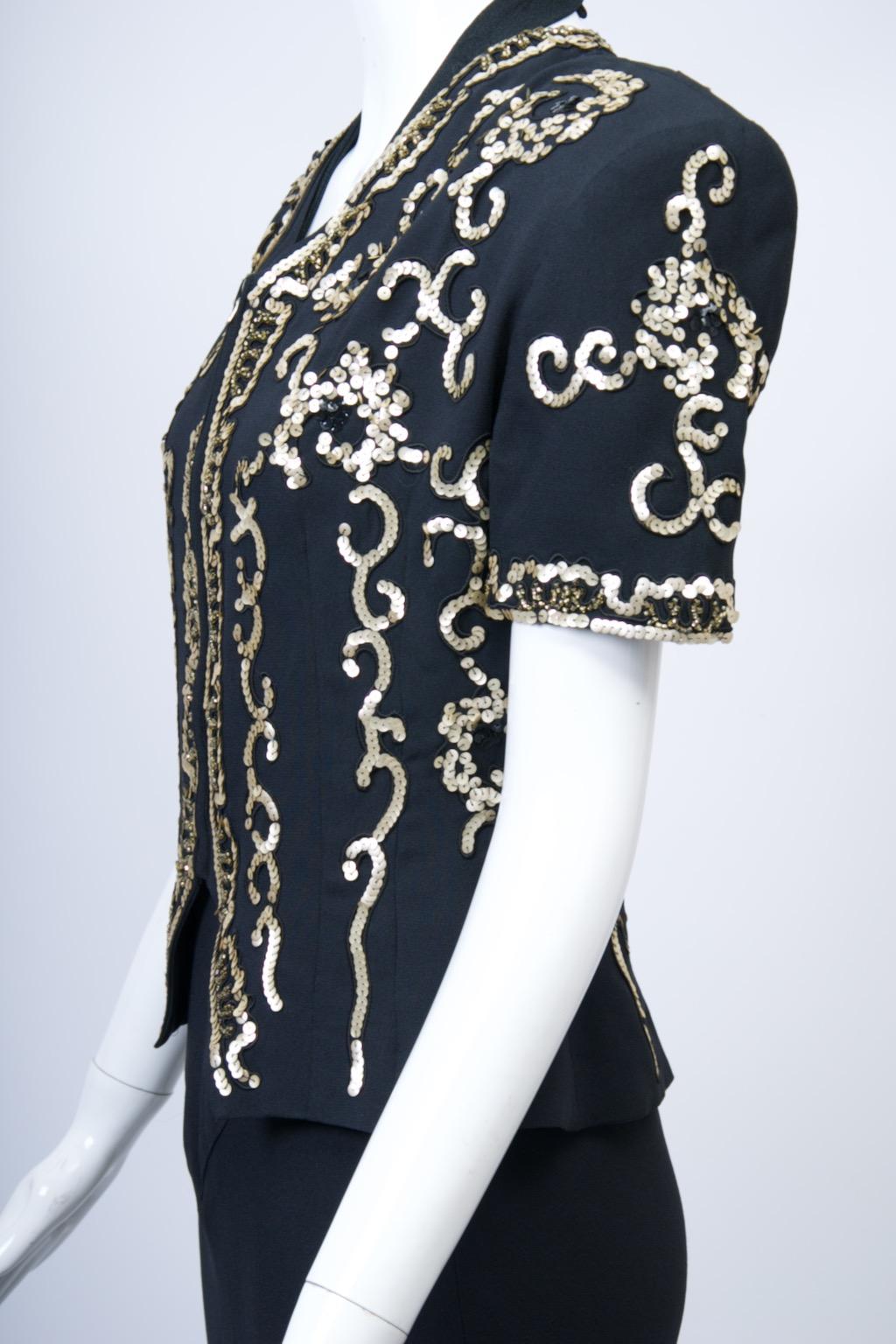 Giacca da sera vintage di Norma Kamali in crêpe nero impreziosita da paillettes dorate in un intricato motivo verticale. La giacca presenta maniche corte con pad, una cerniera frontale e una scollatura a V che si specchia sul fondo. La giacca ha una