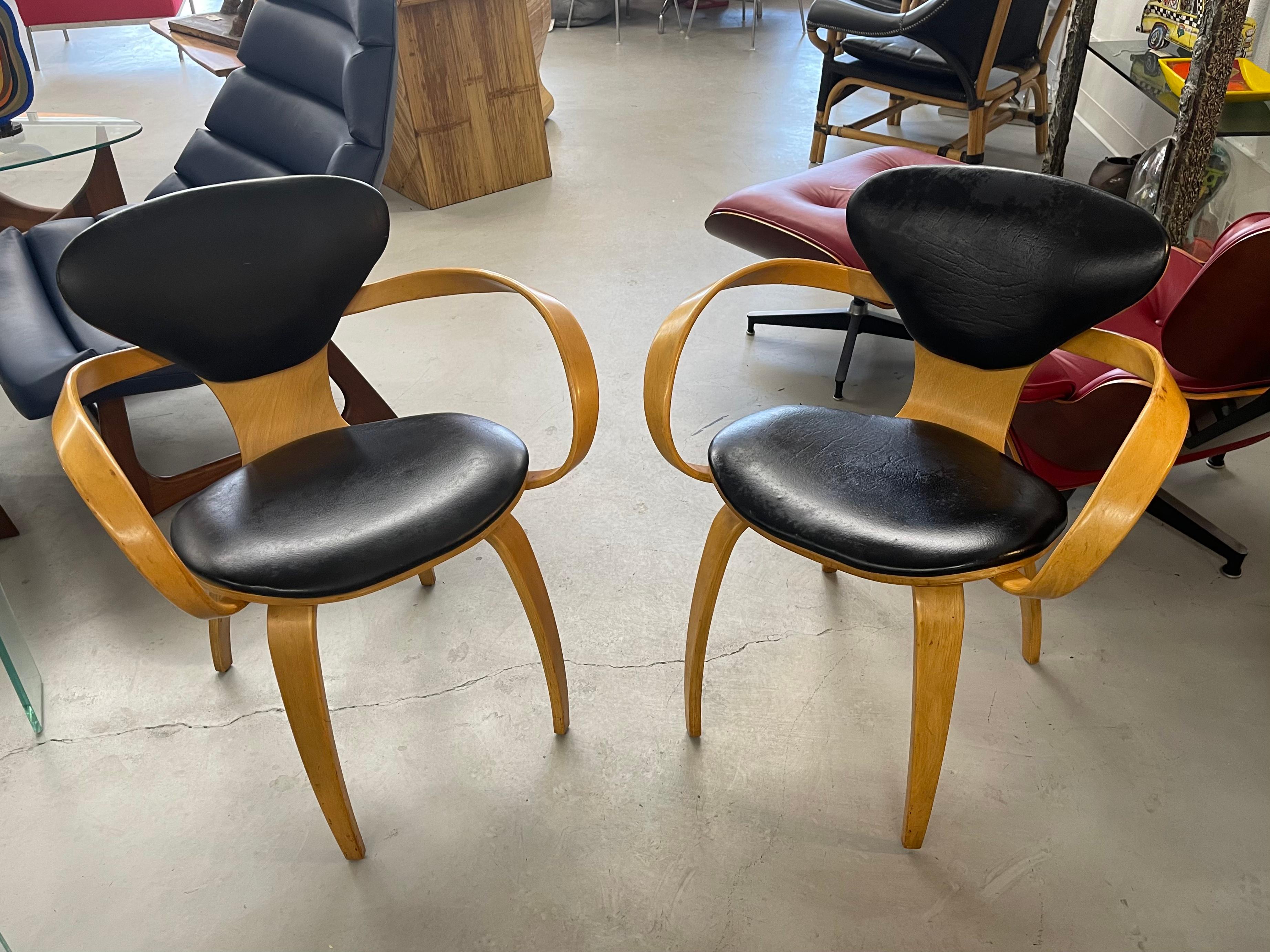 A pair of pretzel armchairs designed by Norman Cherner for Plycraft. These chairs likely date from the 1960’s. Black vinyl cushions. Wood is in good condition with age marks and wear. The vinyl is good but there is some wear and a small imperfection