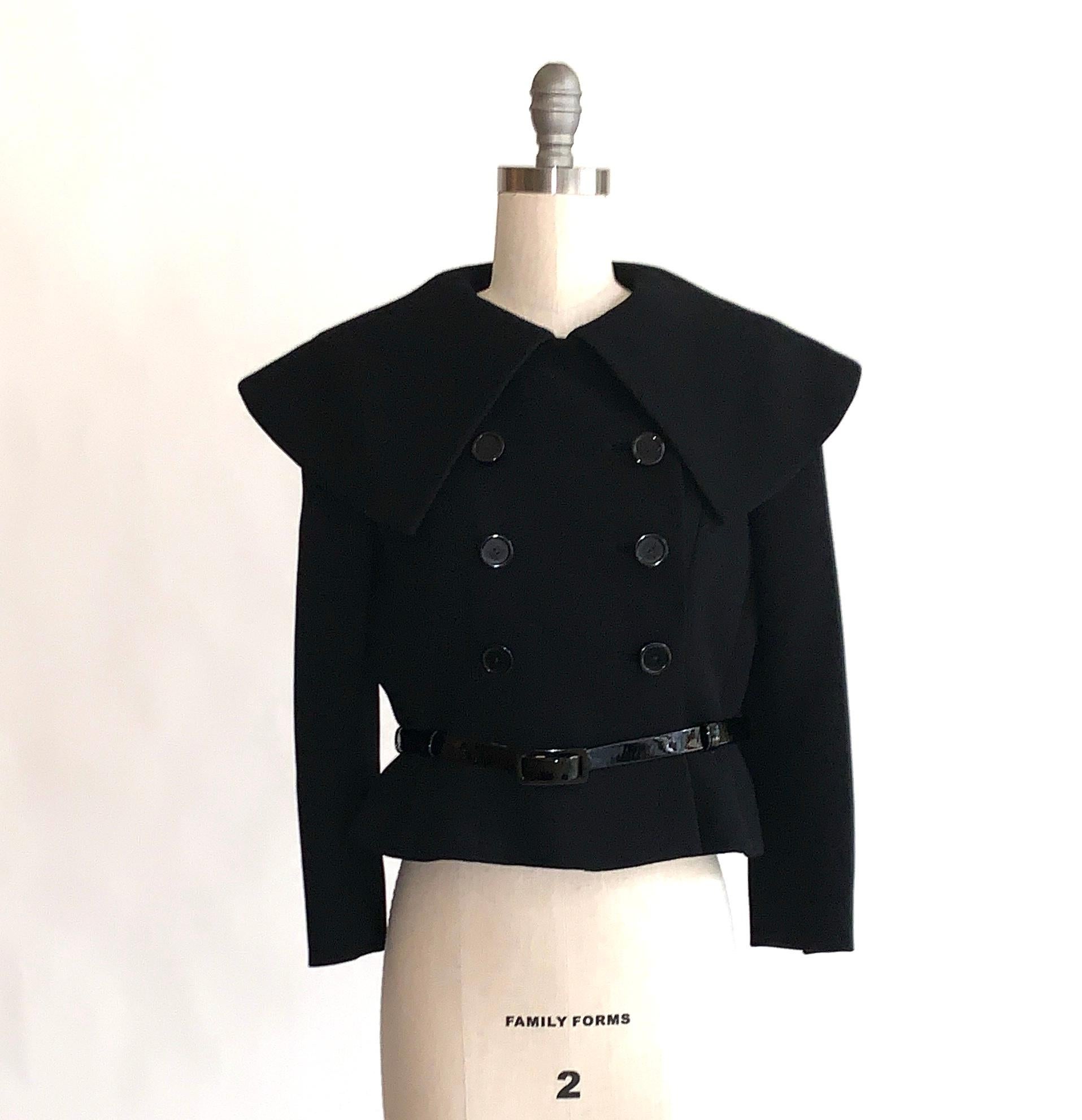 Norman Norell 1960s black belted jacket with oversize pilgrim collar, princess seams, and double breasted button front. Six large plastic buttons at front and three smaller buttons at each cuff. Three belt loops at waist hold a thin patent leather